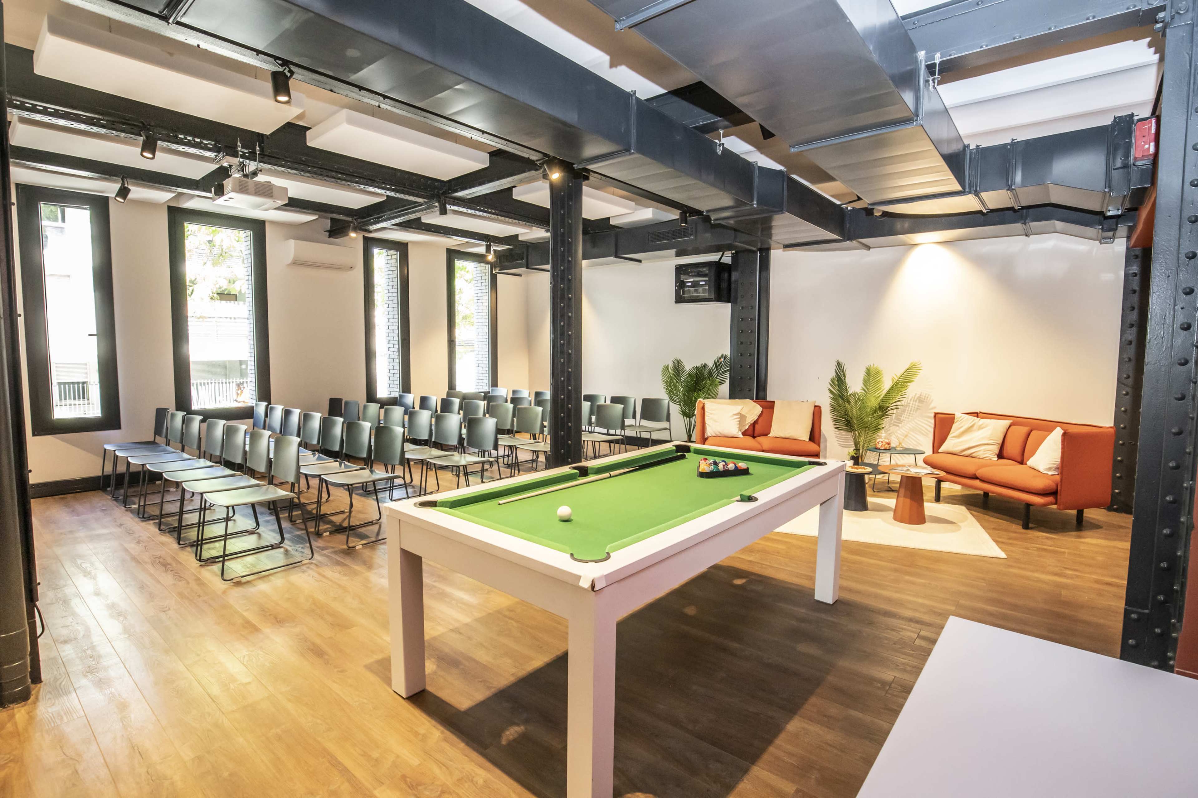 A spacious room features a green pool table centered among rows of metal chairs and a cozy seating area with two armchairs and a coffee table.