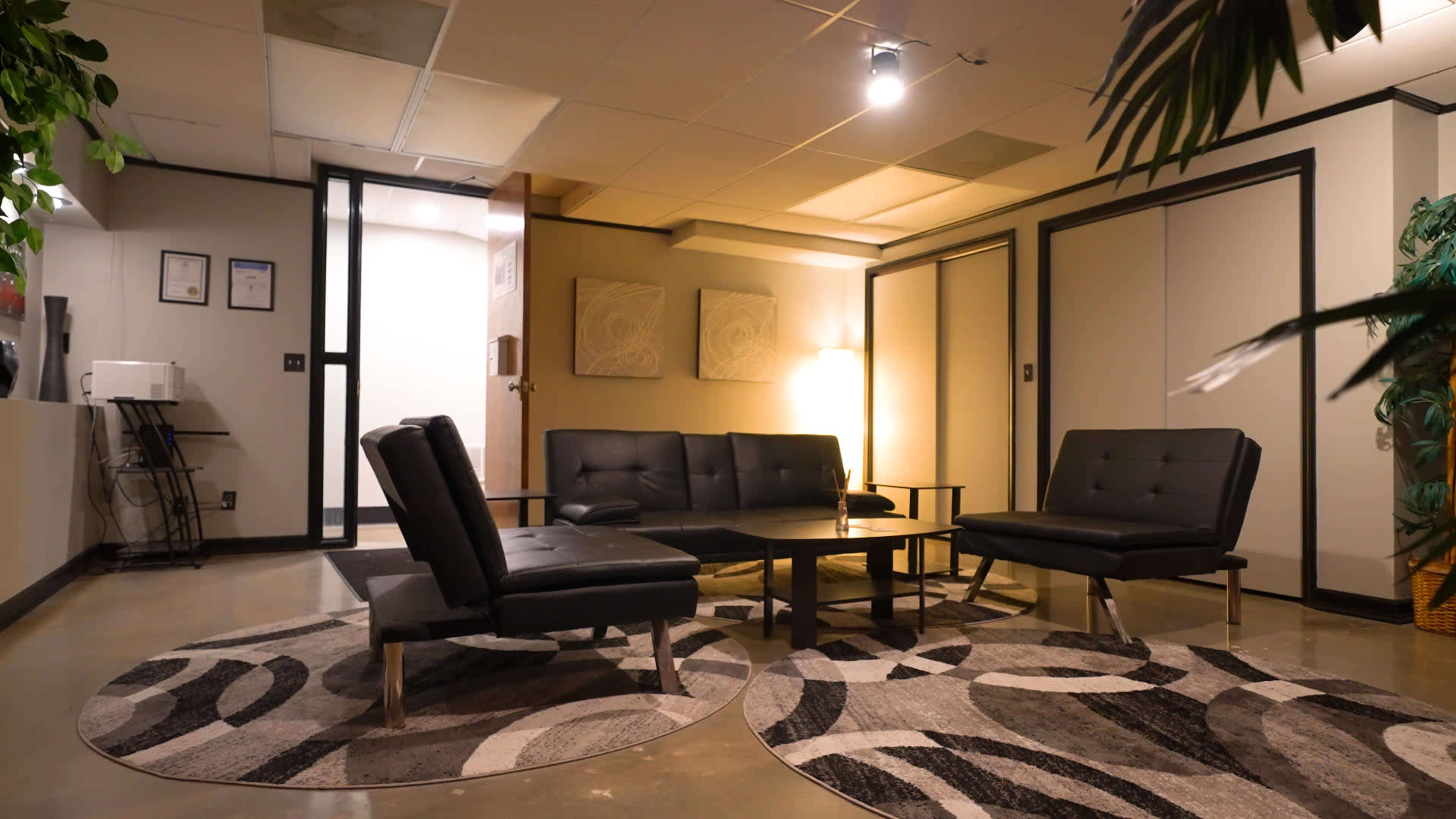 A modern waiting area with black leather furniture, a round coffee table, and patterned area rugs, illuminated by overhead lighting.