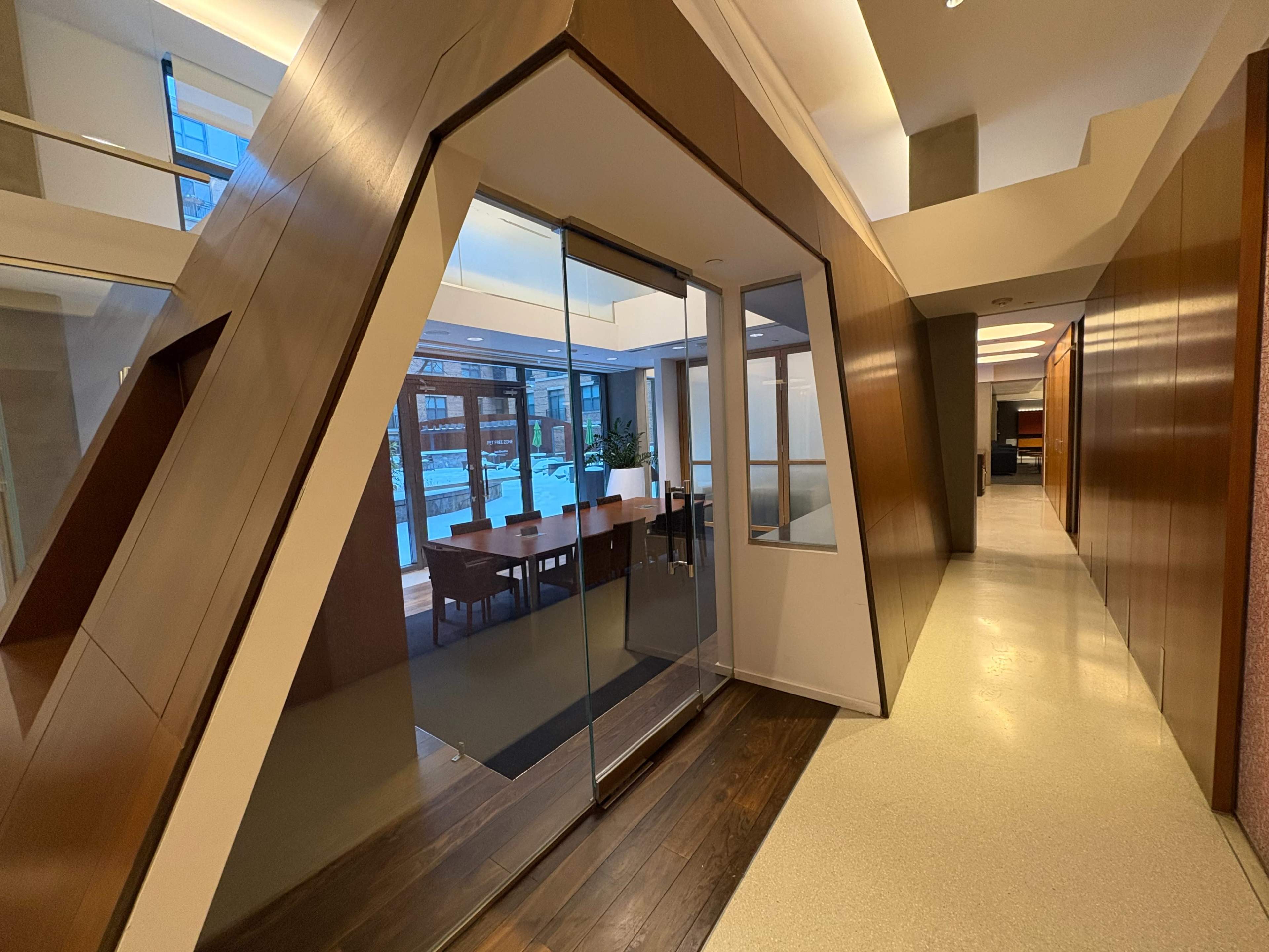 The image shows a modern indoor space featuring a glass-walled meeting room with wooden accents and a view of a table and chairs inside.