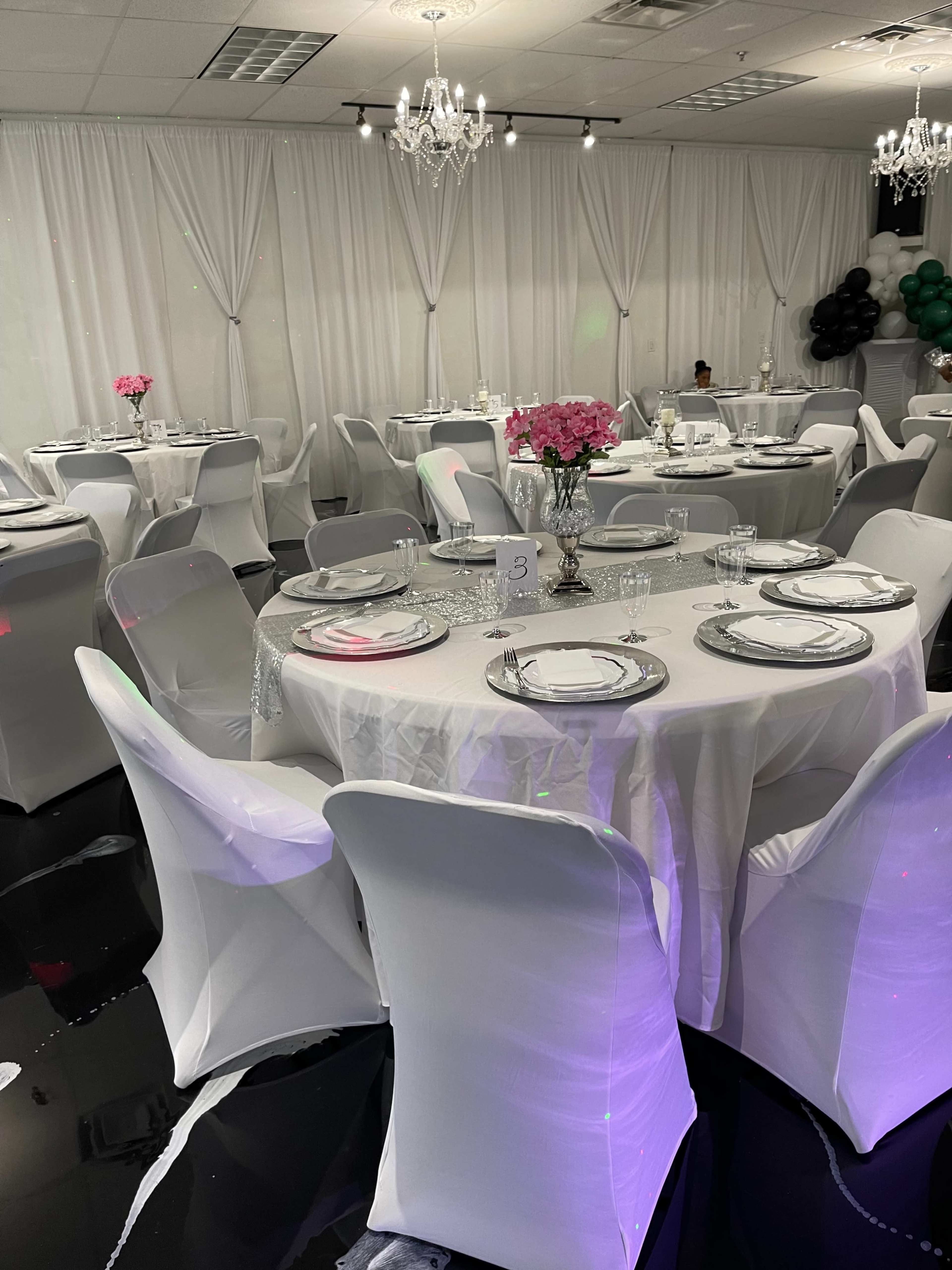 The image shows a banquet room set with elegantly dressed tables, featuring white tablecloths and pink floral centerpieces, under decorative lighting.