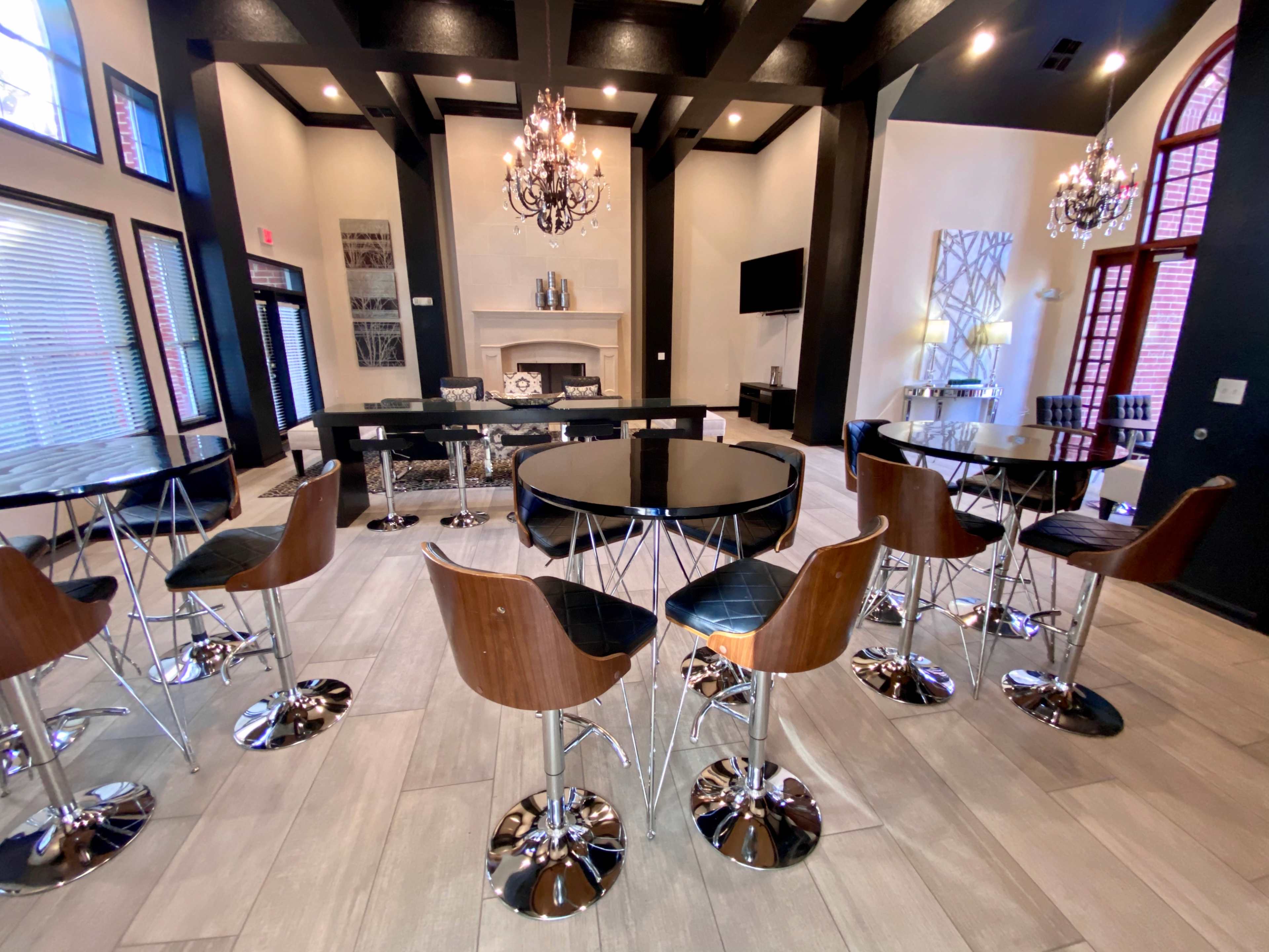 The image shows a contemporary lounge area featuring several round tables surrounded by high-backed chairs, with a fireplace and television in the background.