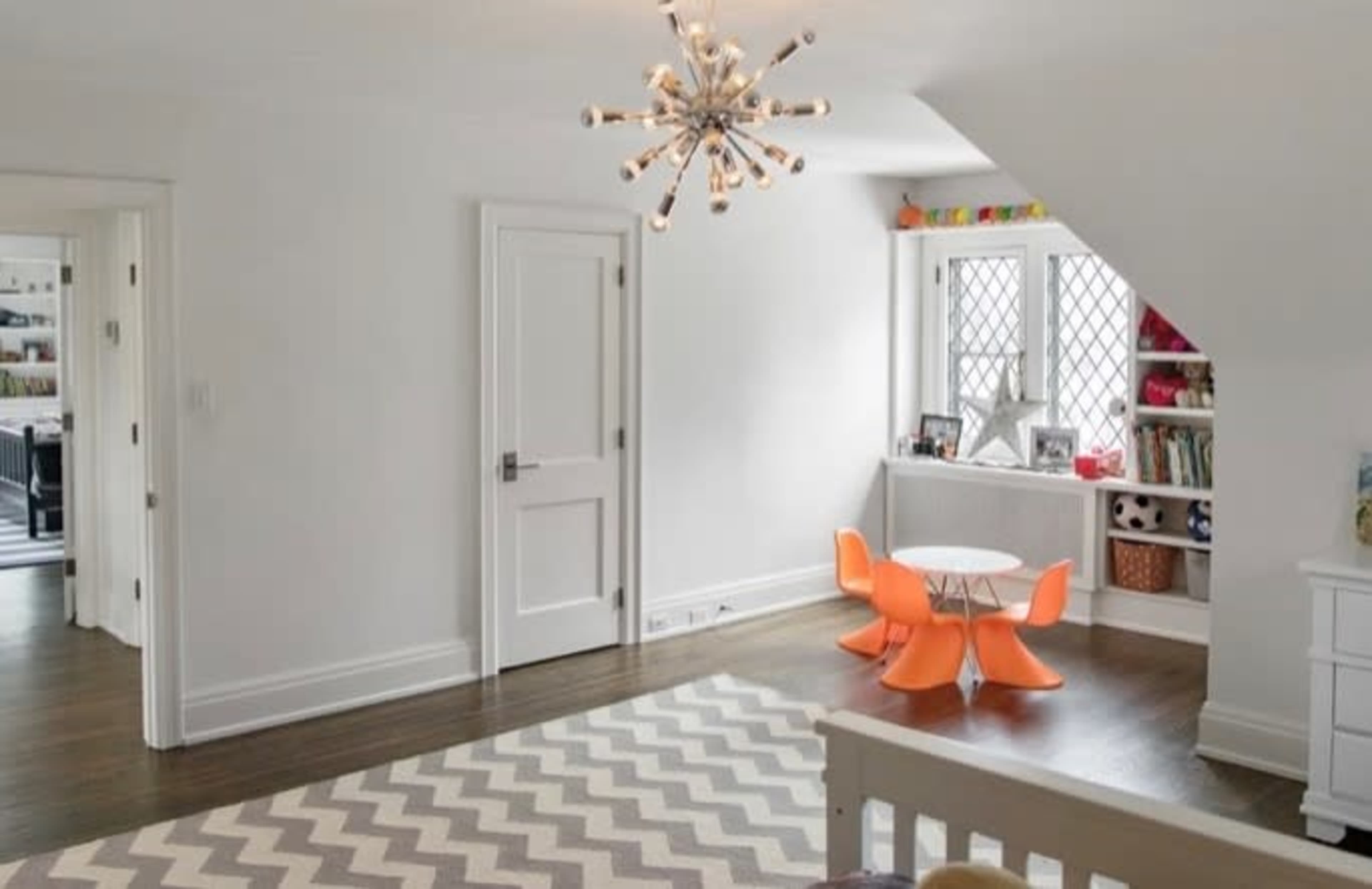 A bright, spacious children's room featuring a round table with orange chairs, a bookshelf, and a striped carpet.