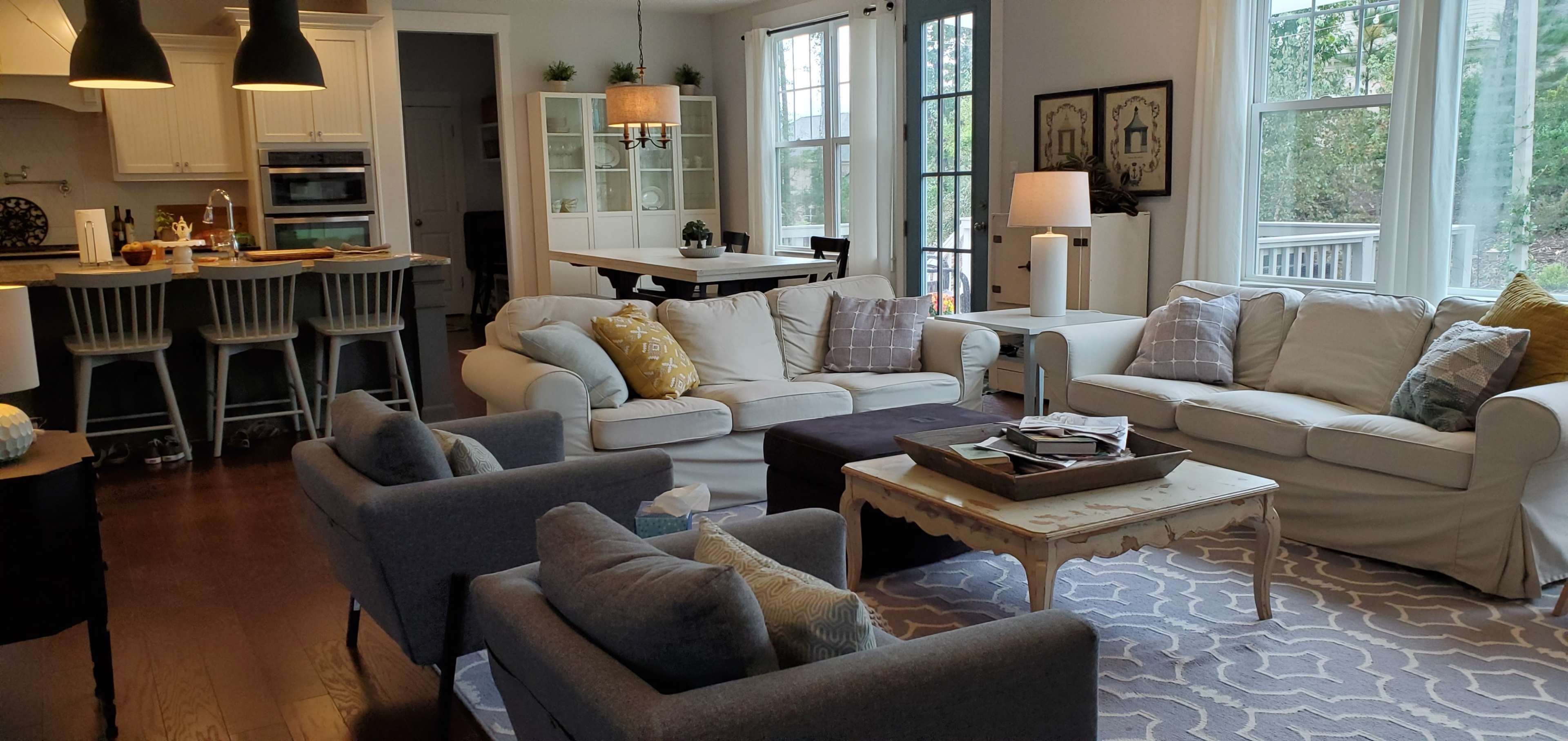 The image shows a modern living room with white sofas, two gray armchairs, and a light rug, connected to a dining area with a table and chairs.