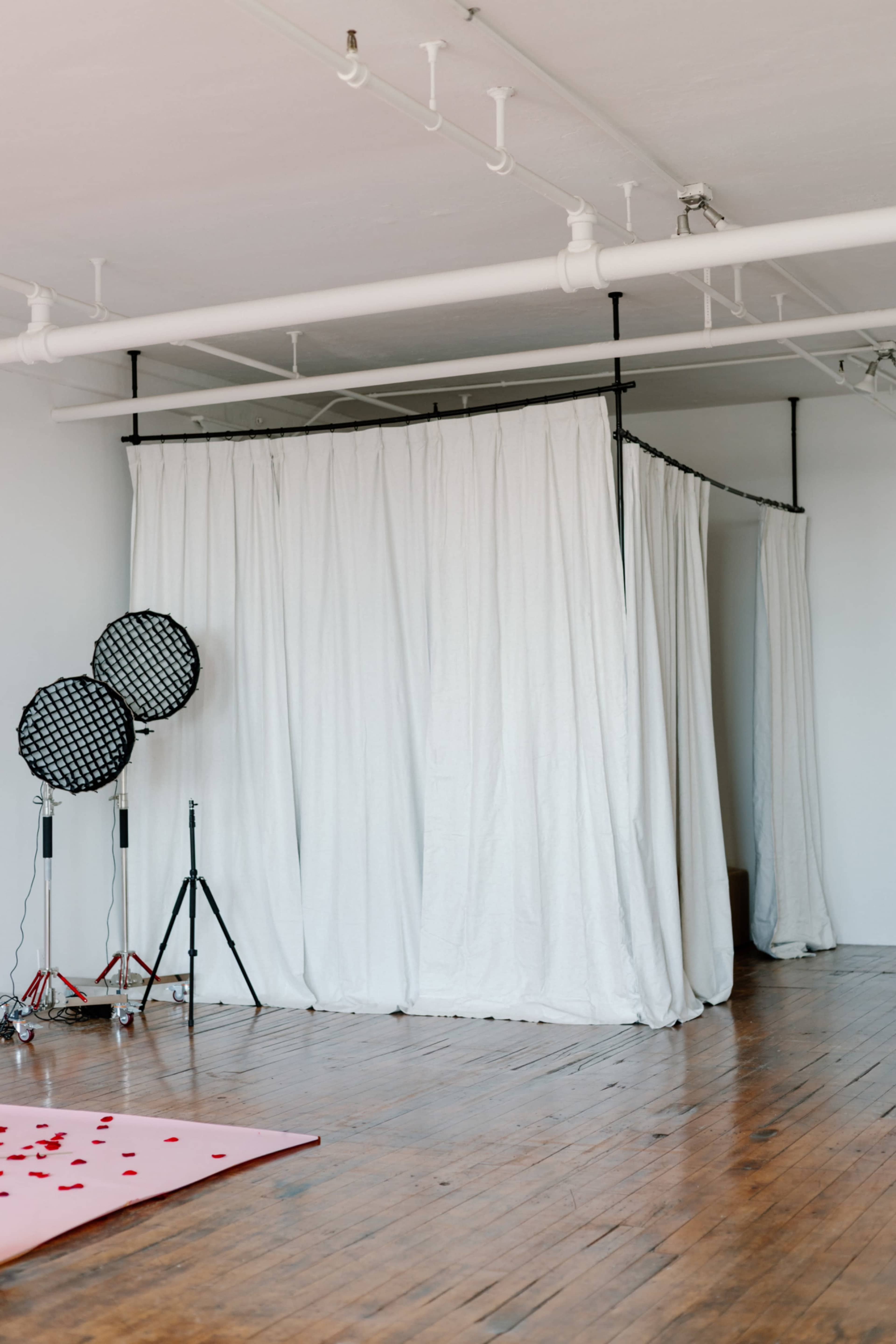 The image shows a spacious room with wooden flooring, featuring a white curtain setup and two blacklights on stands.