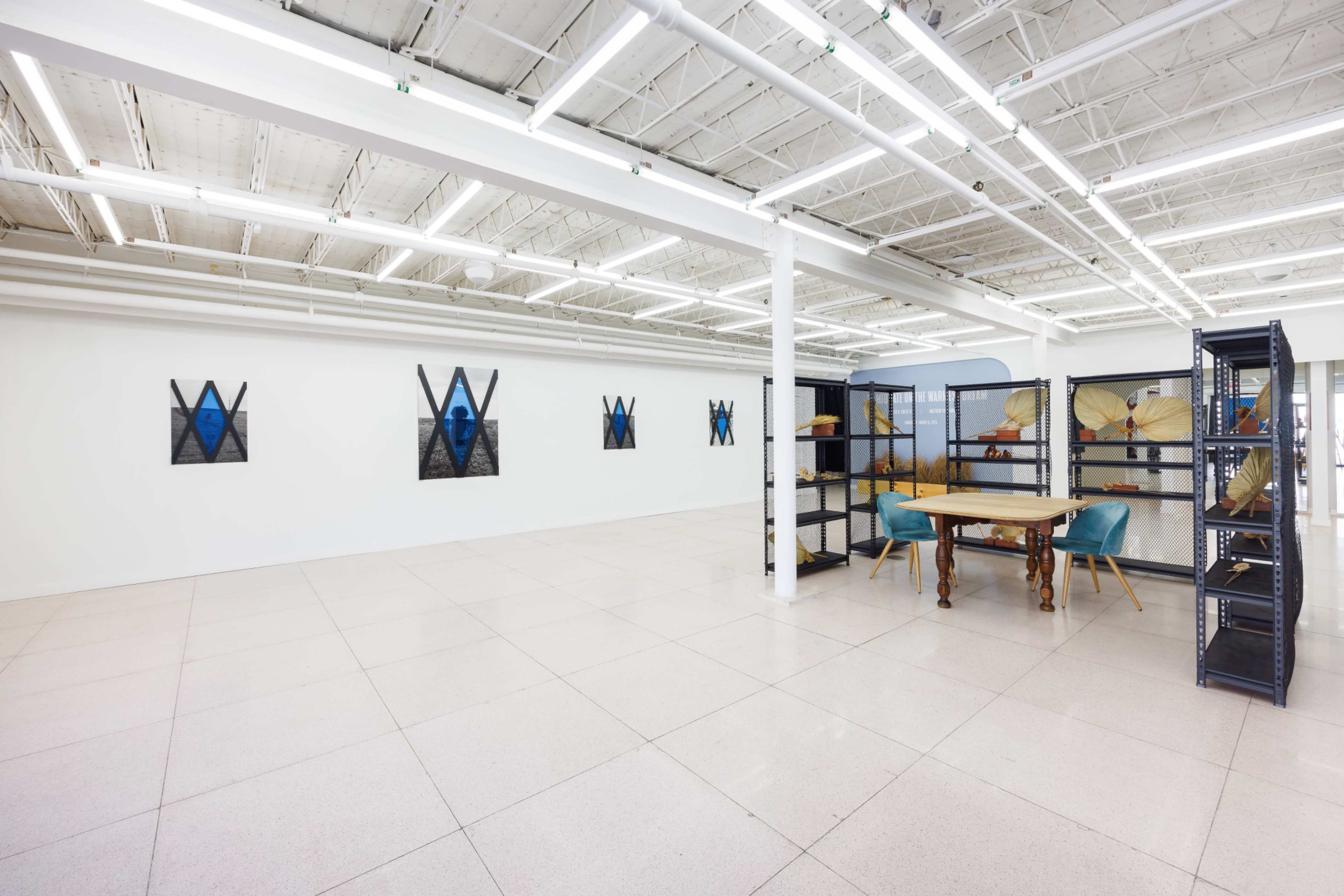 The image shows a spacious, well-lit interior with white walls, featuring artwork on display and a table with two blue chairs next to a shelving unit.