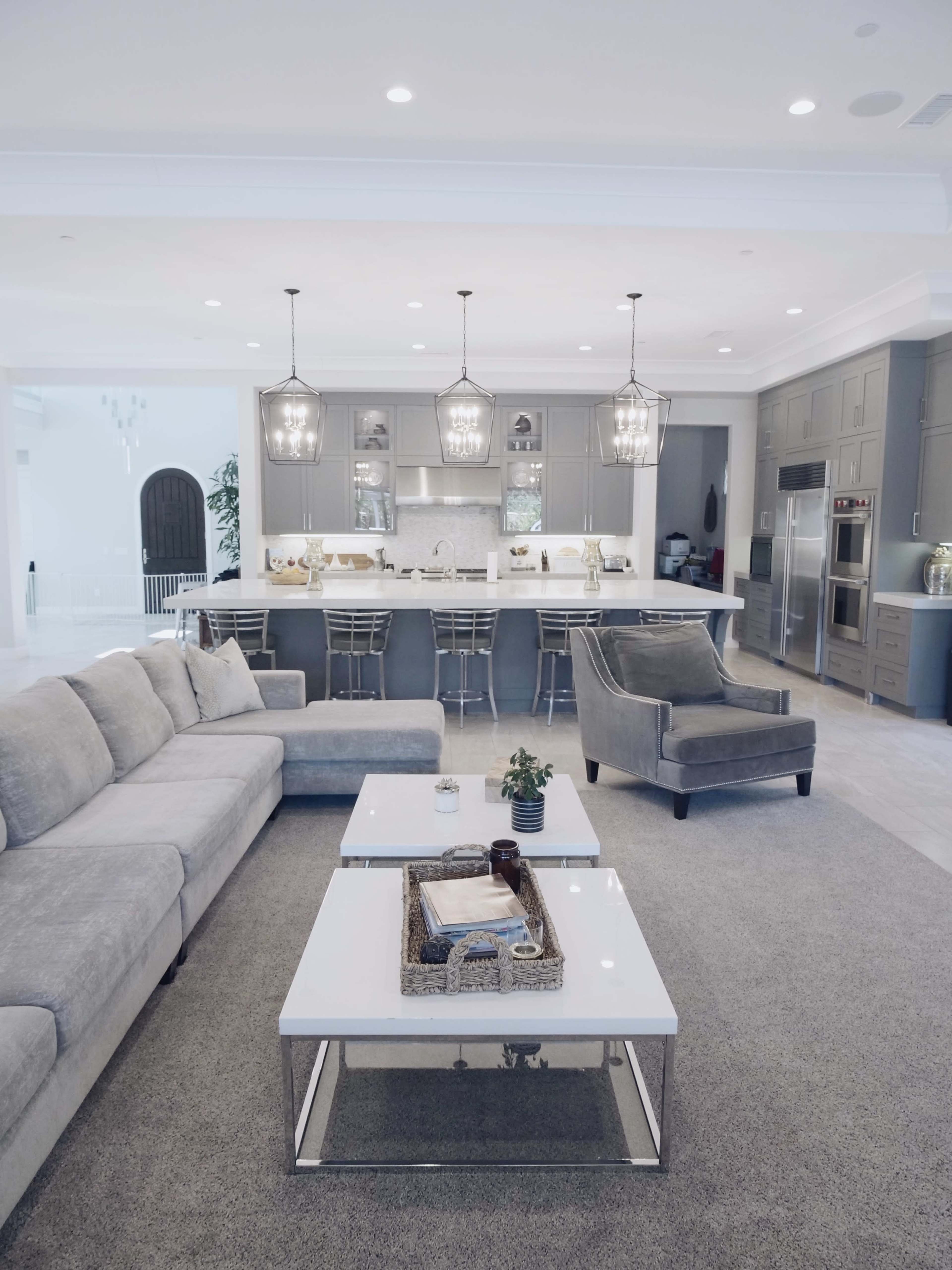 A modern kitchen and living room are depicted, featuring a gray sofa, a white coffee table, and gray cabinetry with pendant lighting above the island.