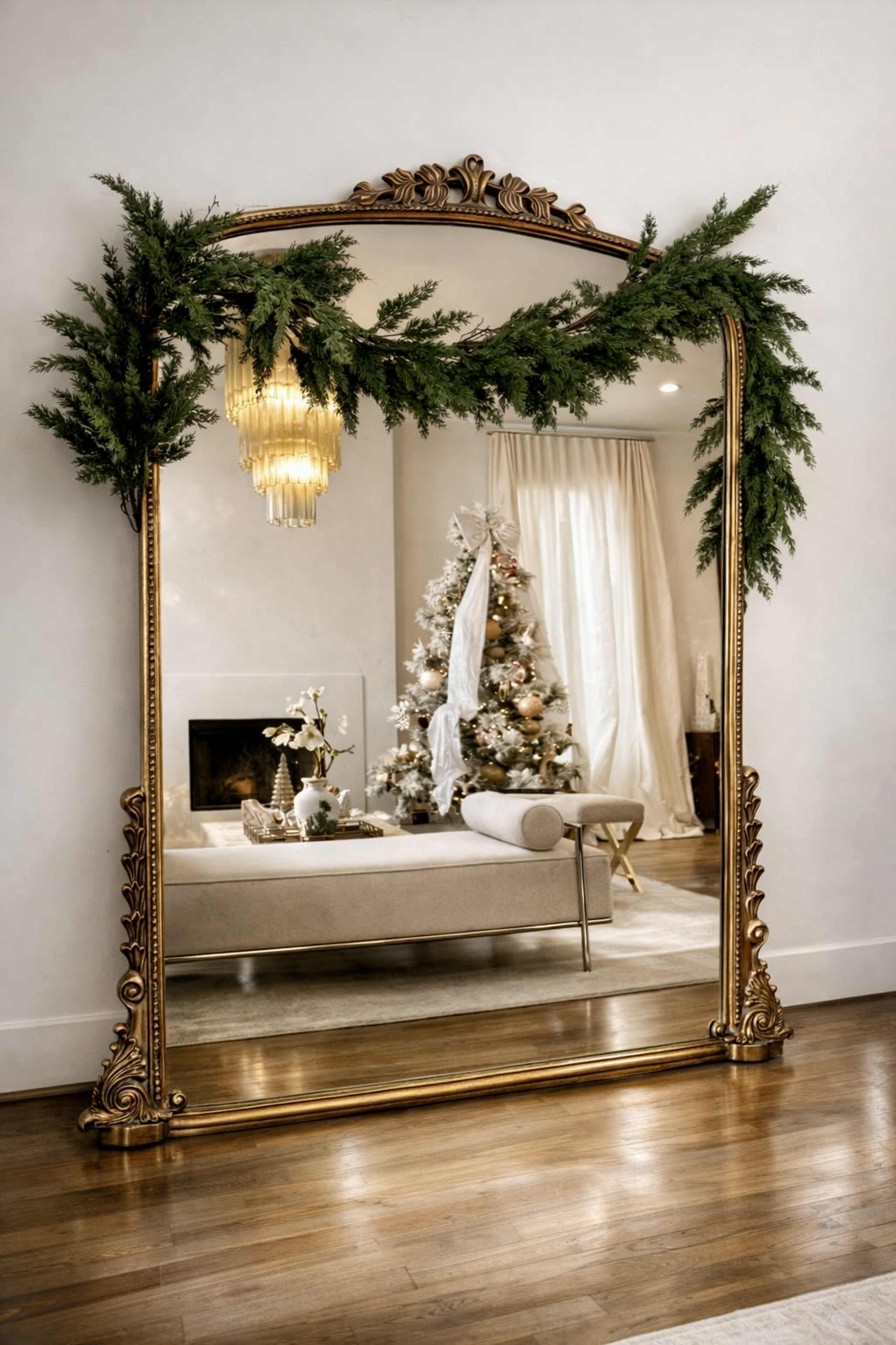 A large, ornate mirror adorned with greenery reflects a cozy living room featuring a decorated Christmas tree and elegant furnishings.