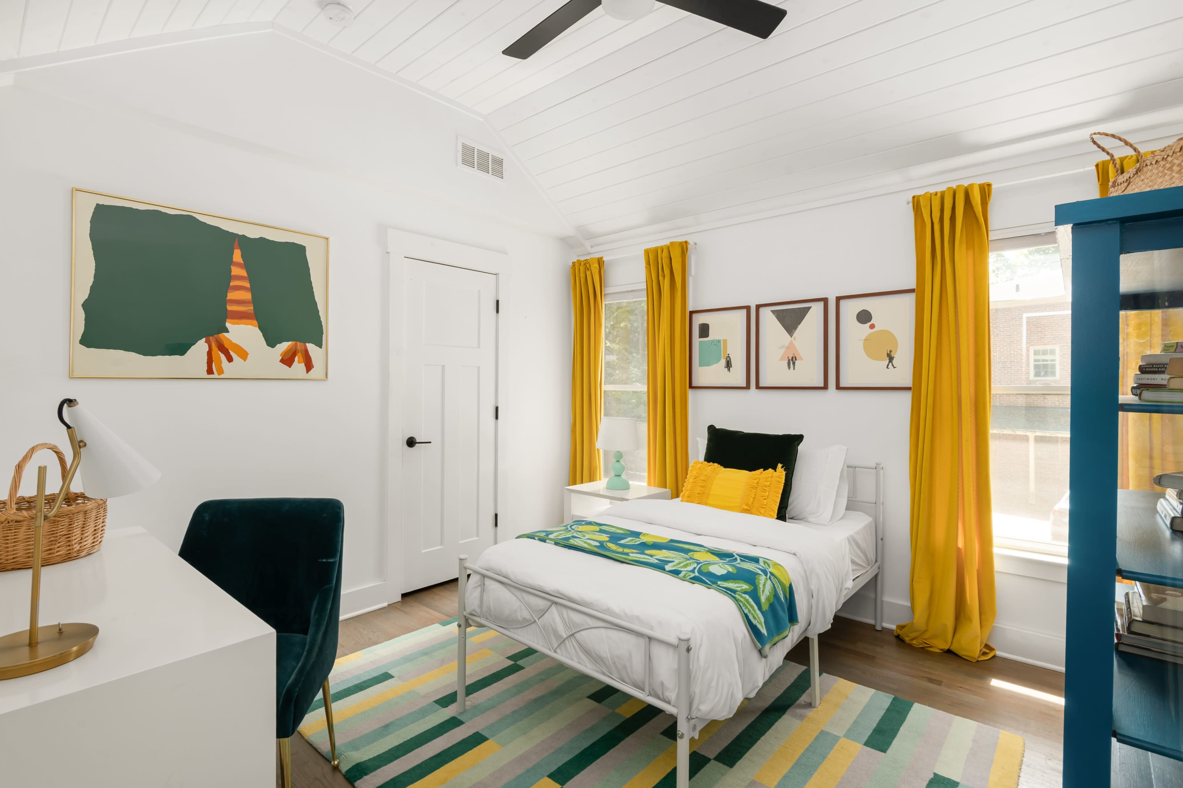 The image shows a bright bedroom featuring a single bed with white bedding, a green accent chair, a desk with a lamp, and colorful artwork and curtains.