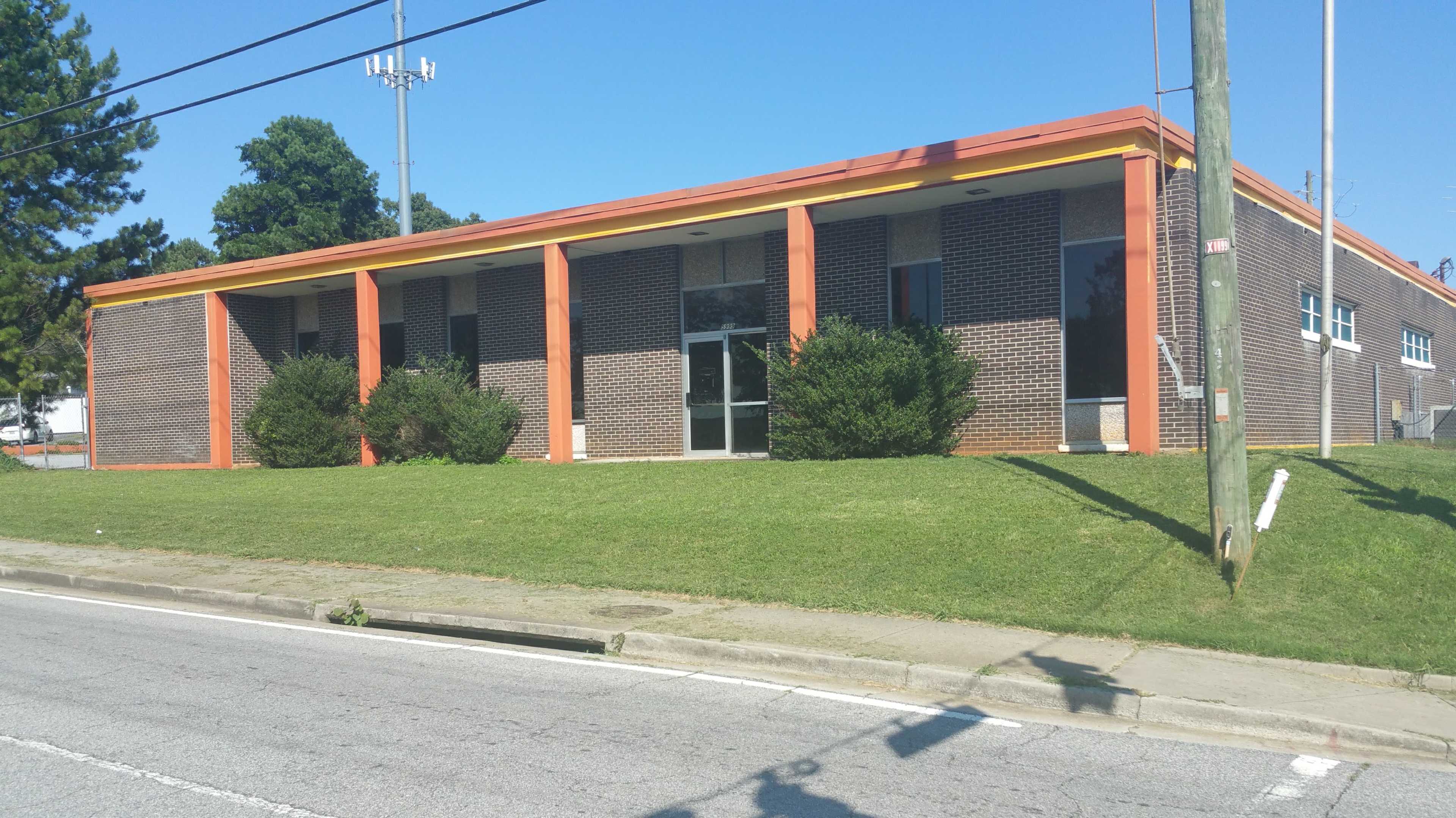 The image shows a single-story brick building with orange trim, situated on a grassy slope along a road.
