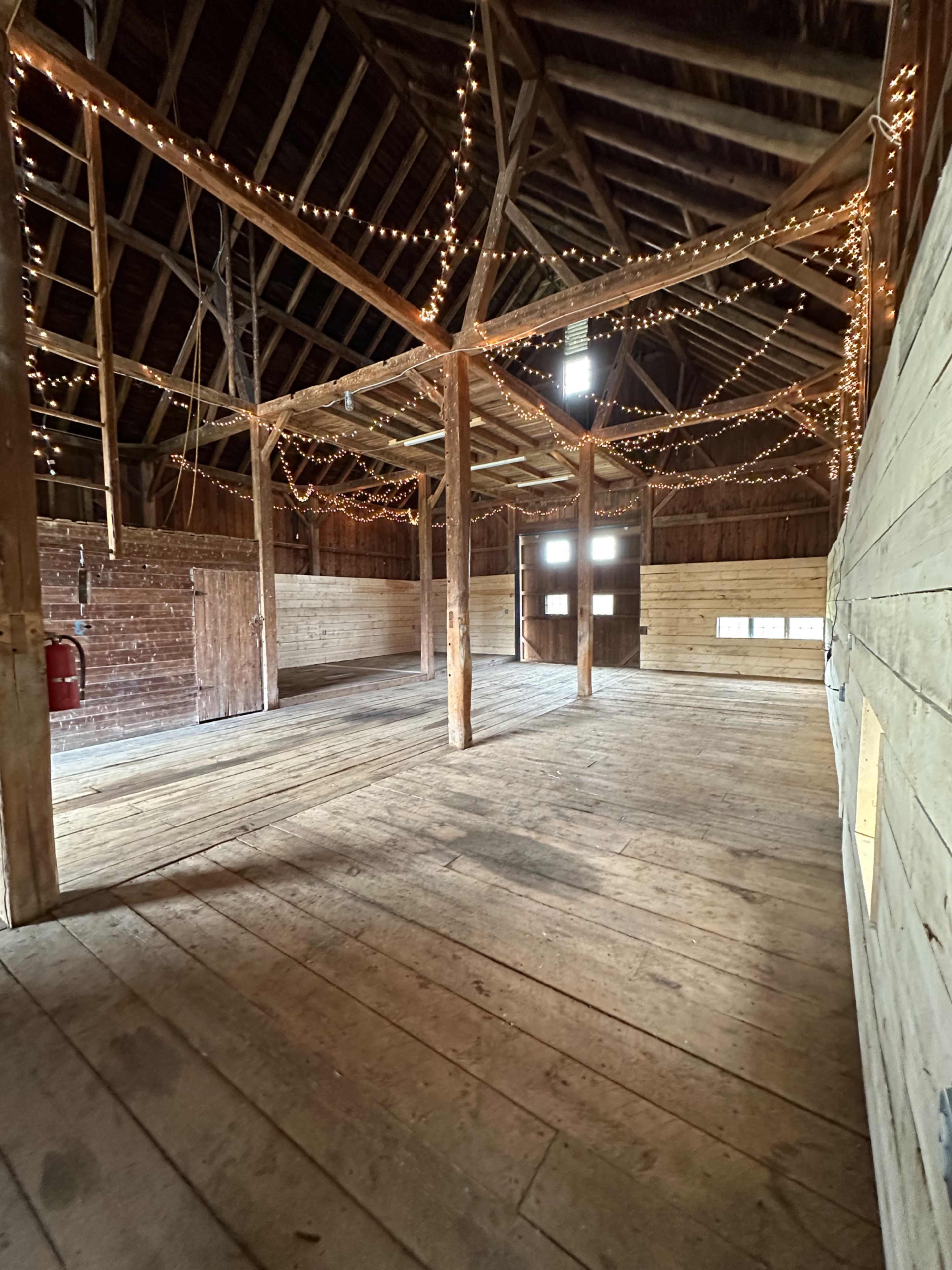 The image shows the interior of a rustic barn, featuring wooden walls, a wooden floor, and string lights hanging from the ceiling beams.