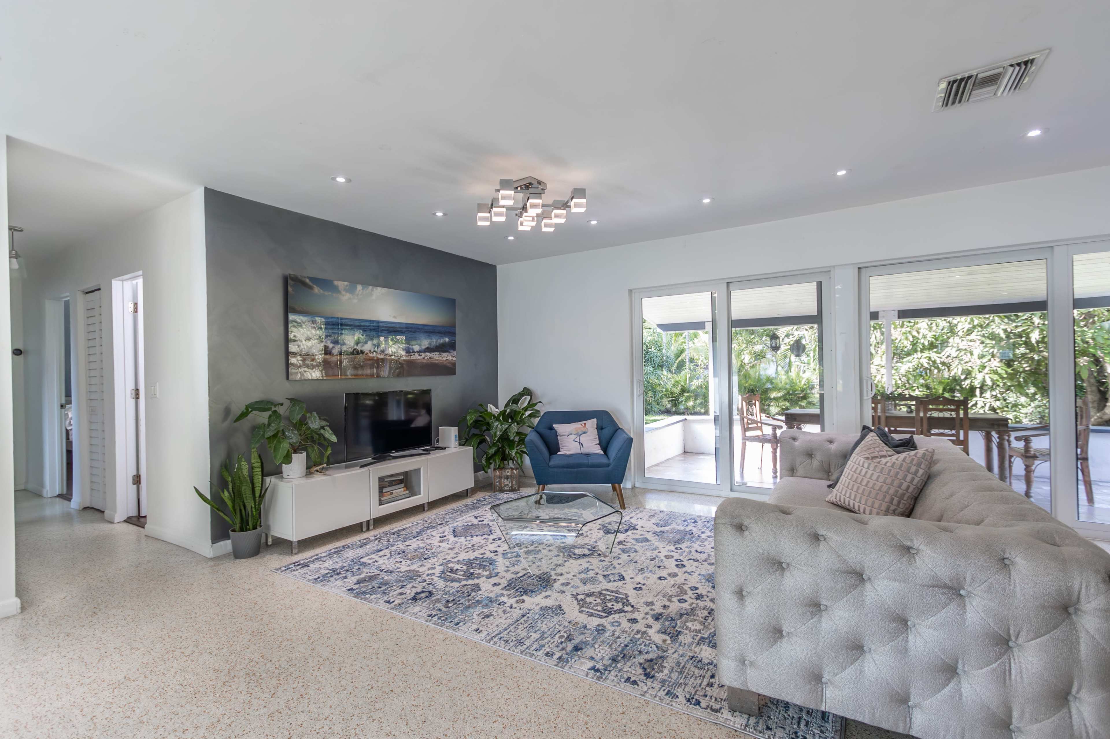 The living room features a blue accent chair, a light grey sofa, a television stand, and large glass doors leading to an outdoor space.