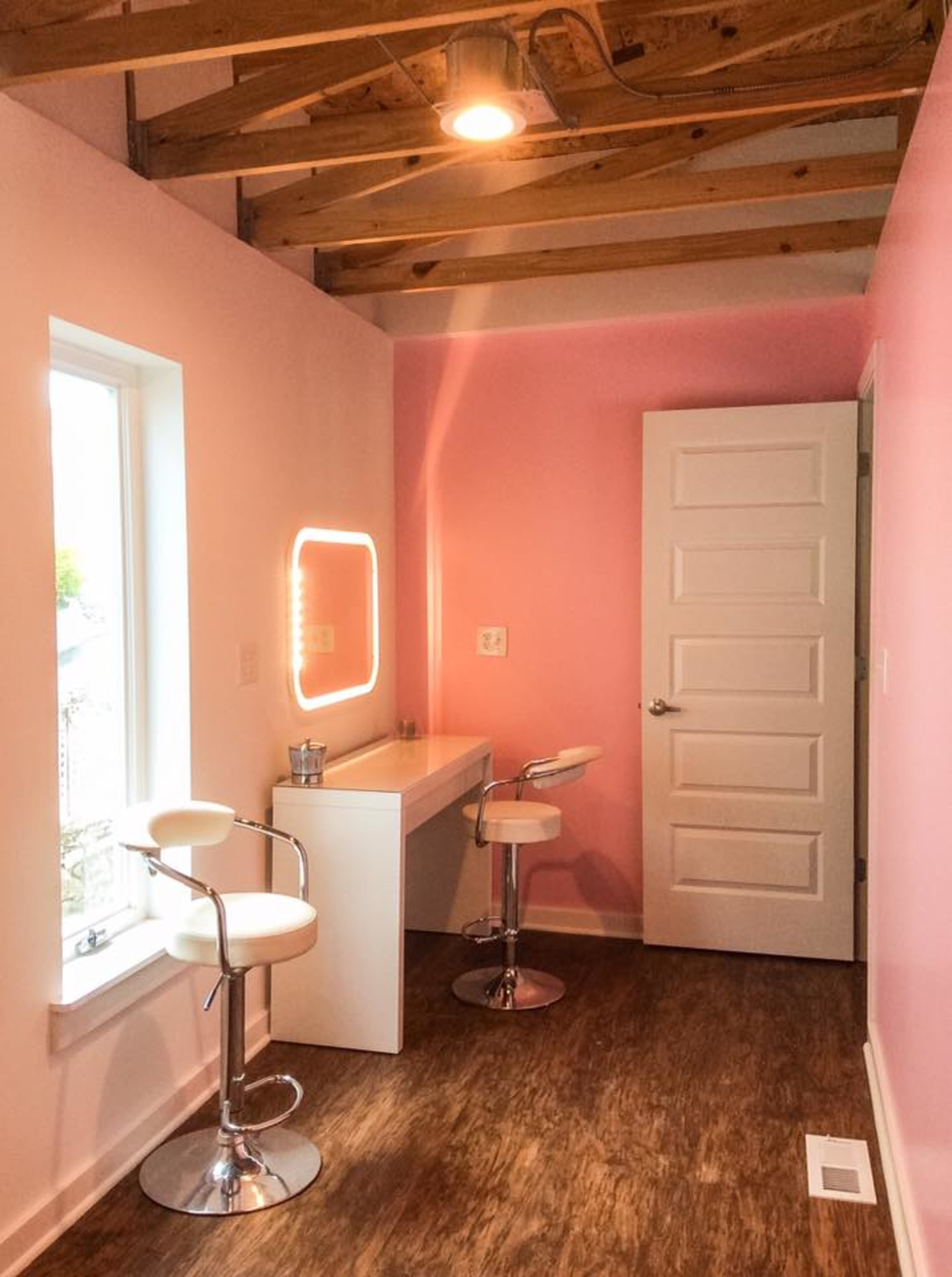 A makeup vanity area with two stools is positioned in a room featuring pink walls and exposed wooden beams.