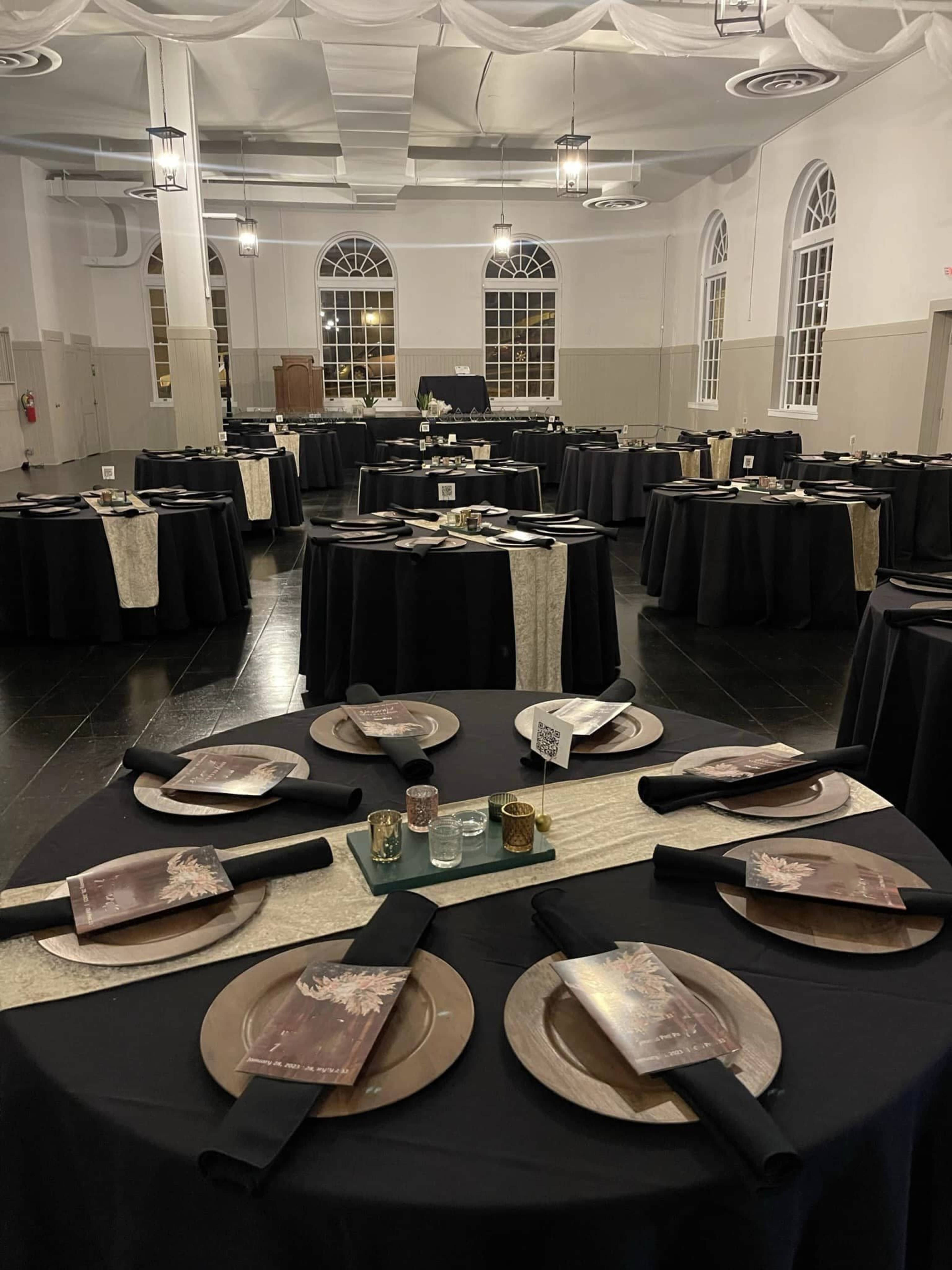 A banquet hall set up with multiple round tables covered in black tablecloths, each decorated with plates, utensils, and folded napkins.