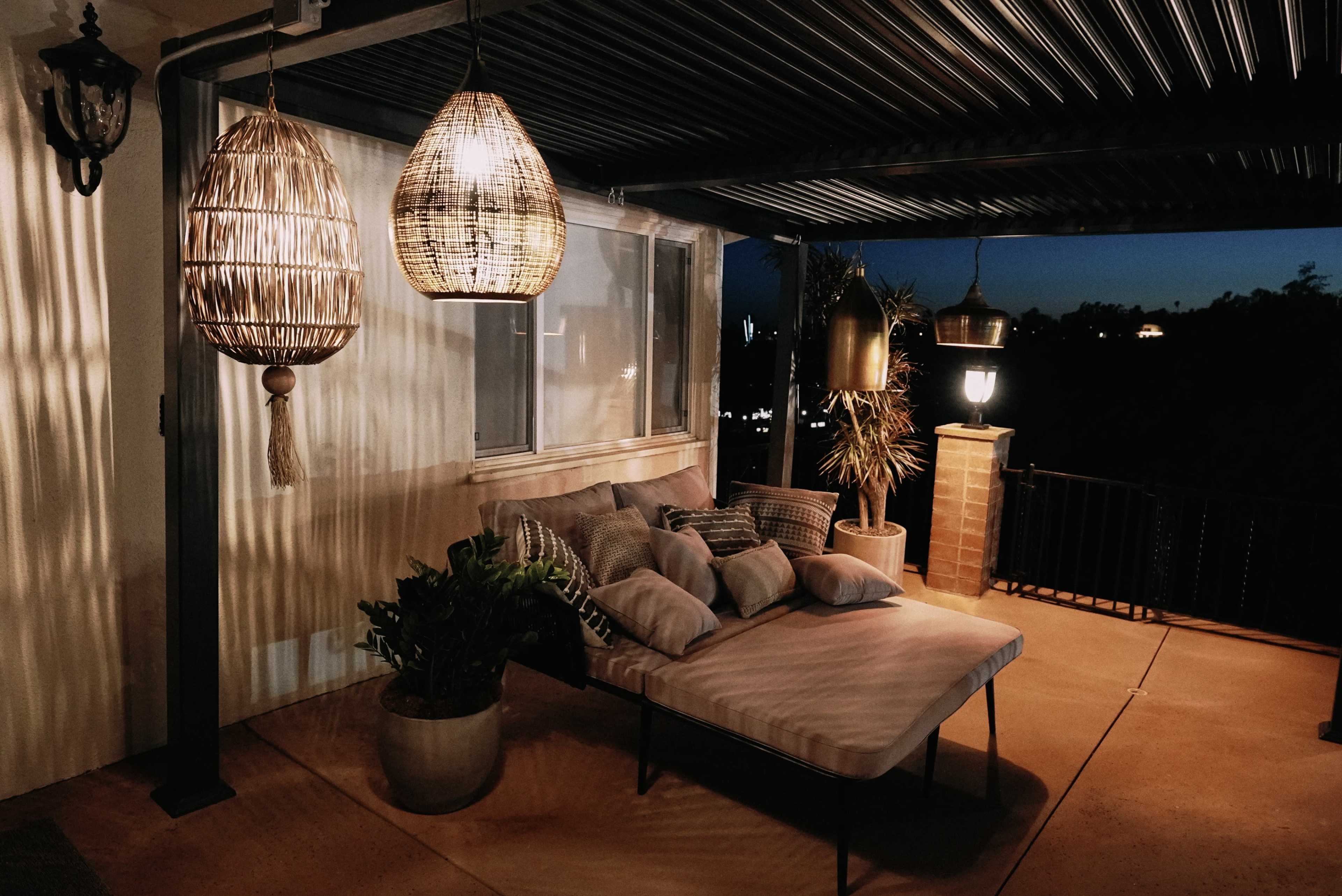 The image shows a cozy outdoor seating area with a sofa adorned with cushions, surrounded by hanging light fixtures and a potted plant.