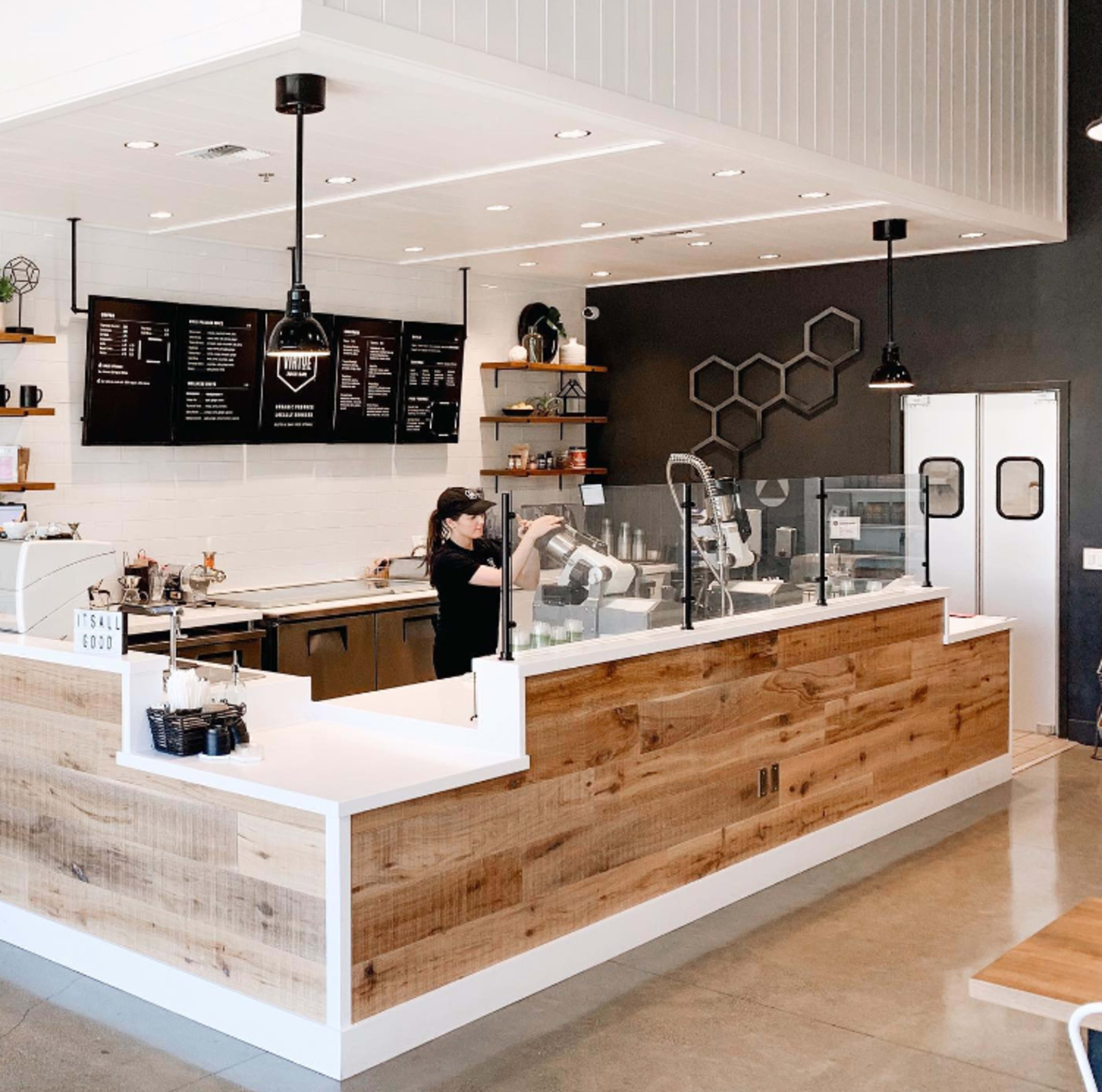 A modern café interior features a wooden serving counter, menu boards on the wall, and a barista preparing drinks.
