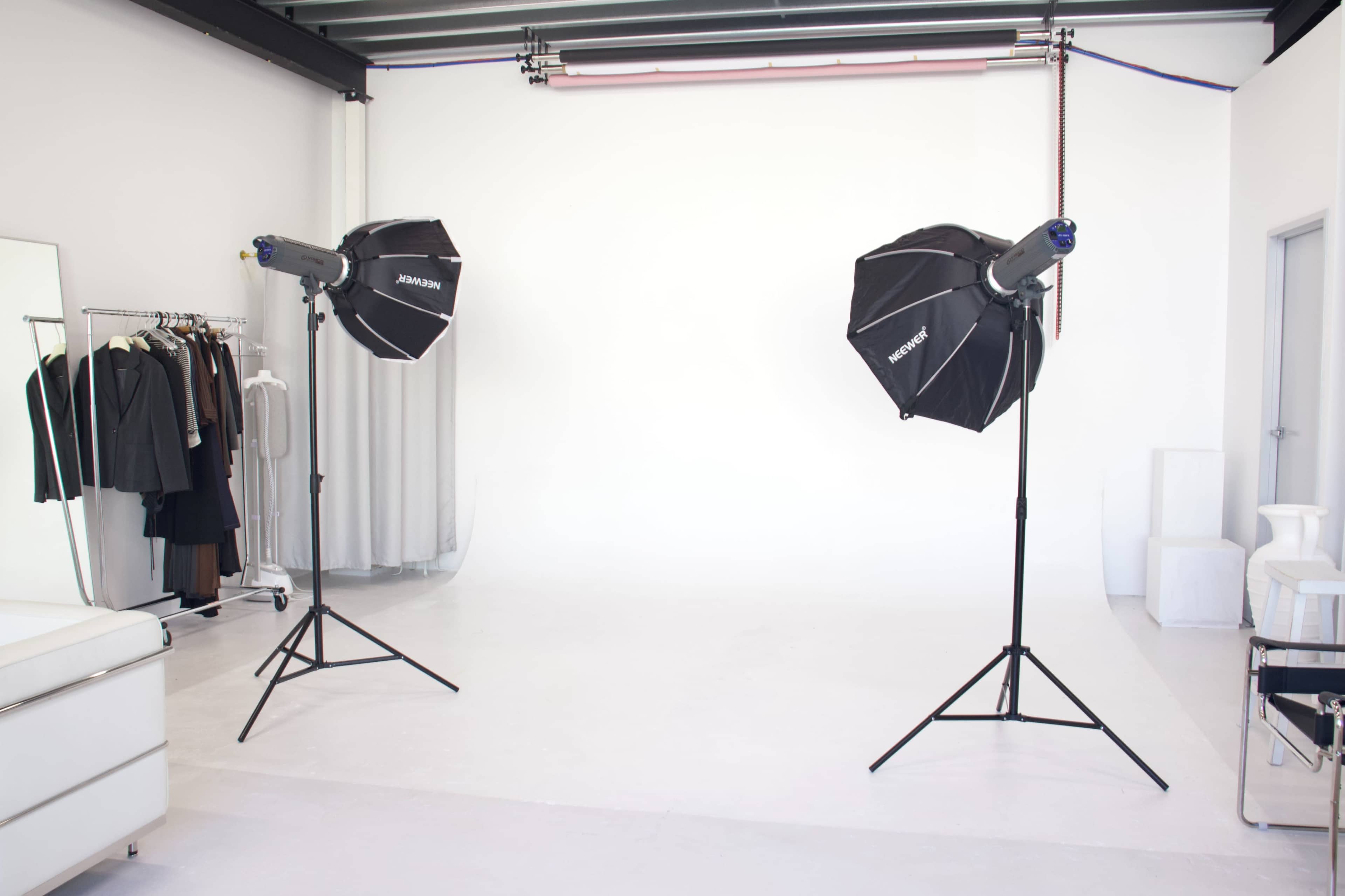 The image shows a photography studio with two softbox lights positioned on stands and a clothing rack featuring various garments against a plain white backdrop.