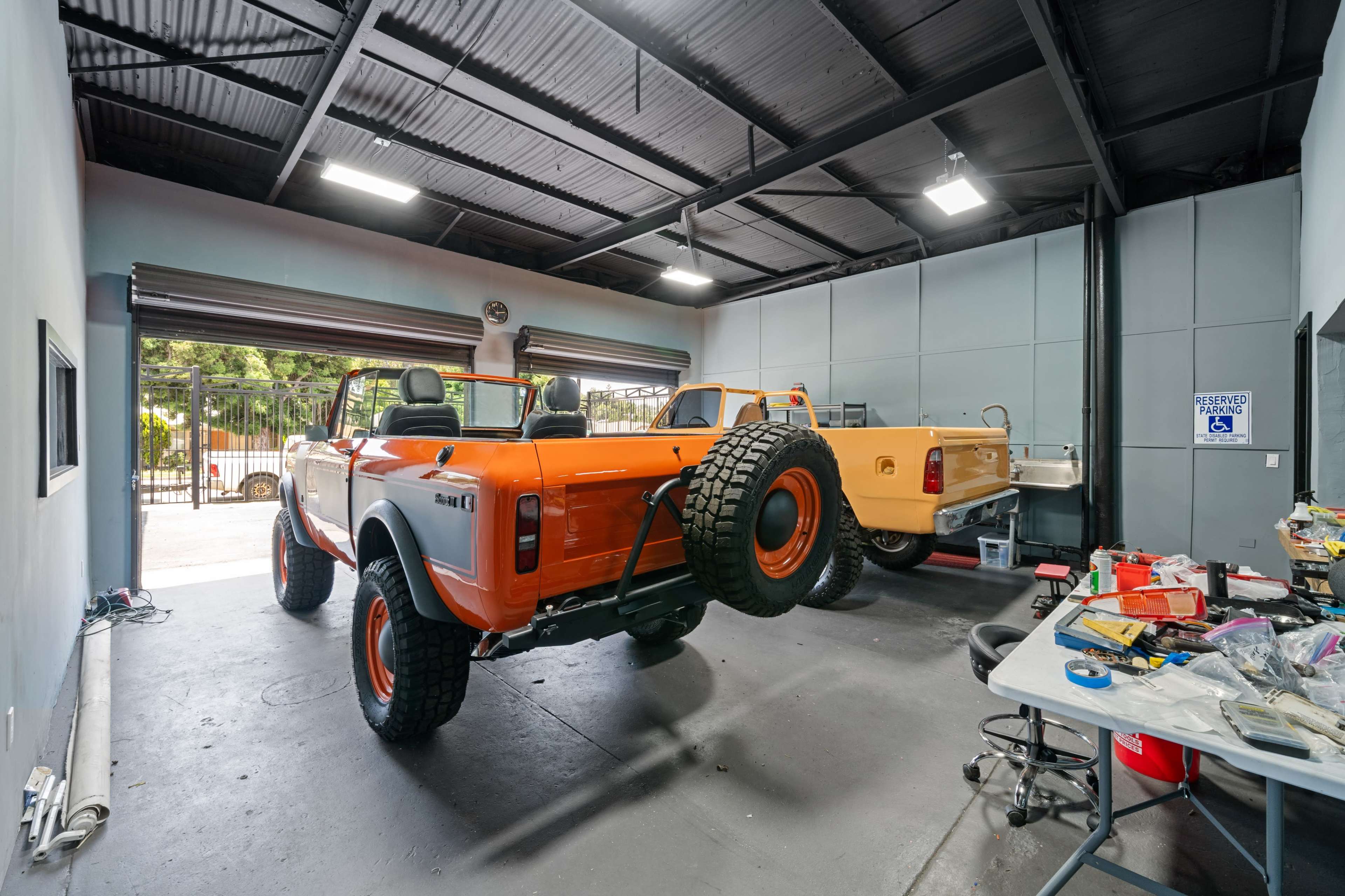 The image shows two vehicles, one orange and one beige, inside a garage with tools and assorted items scattered on the floor.