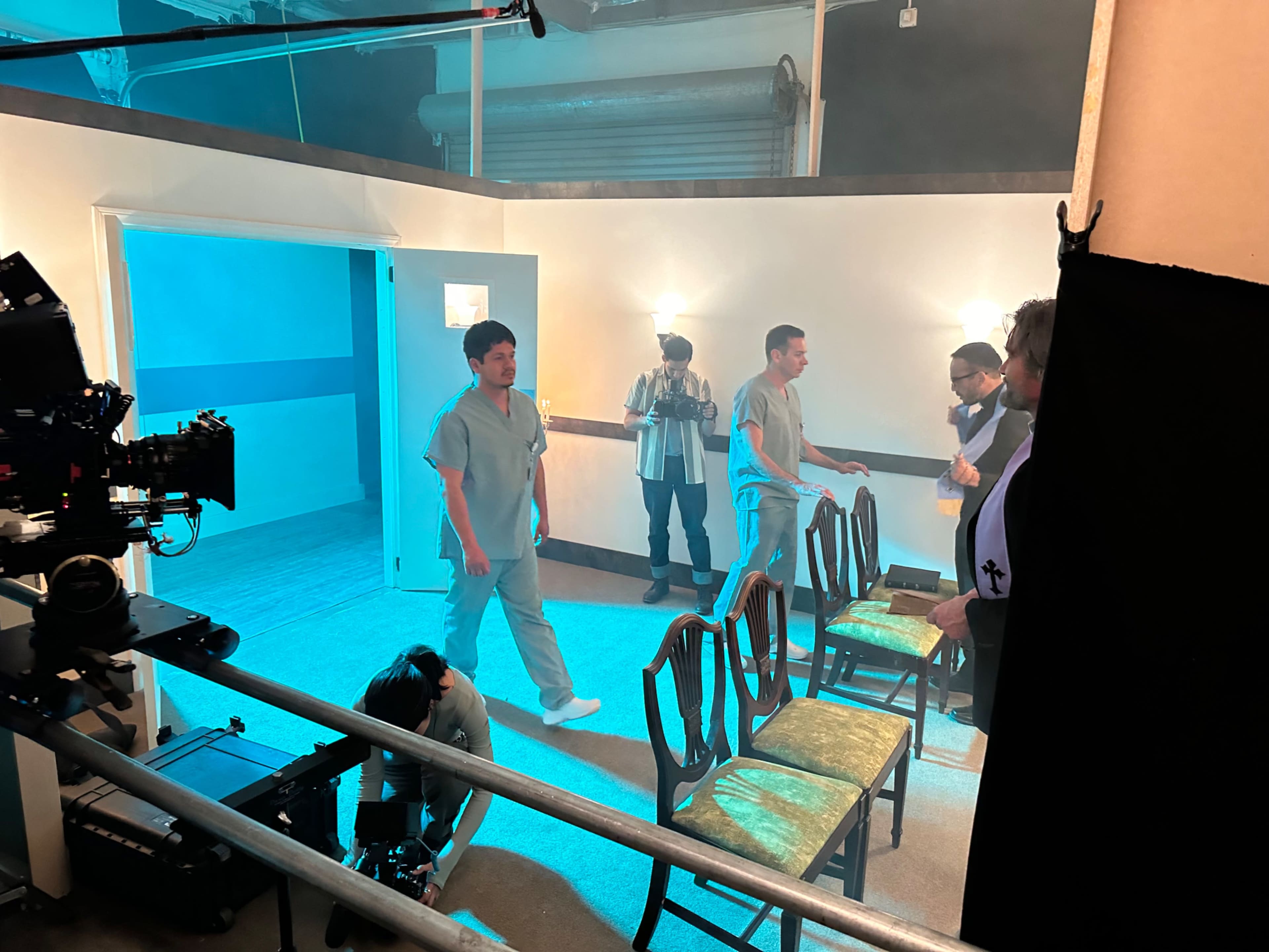 The image shows a film set with medical-themed decorations, featuring multiple actors in scrubs and several empty chairs arranged in the scene.