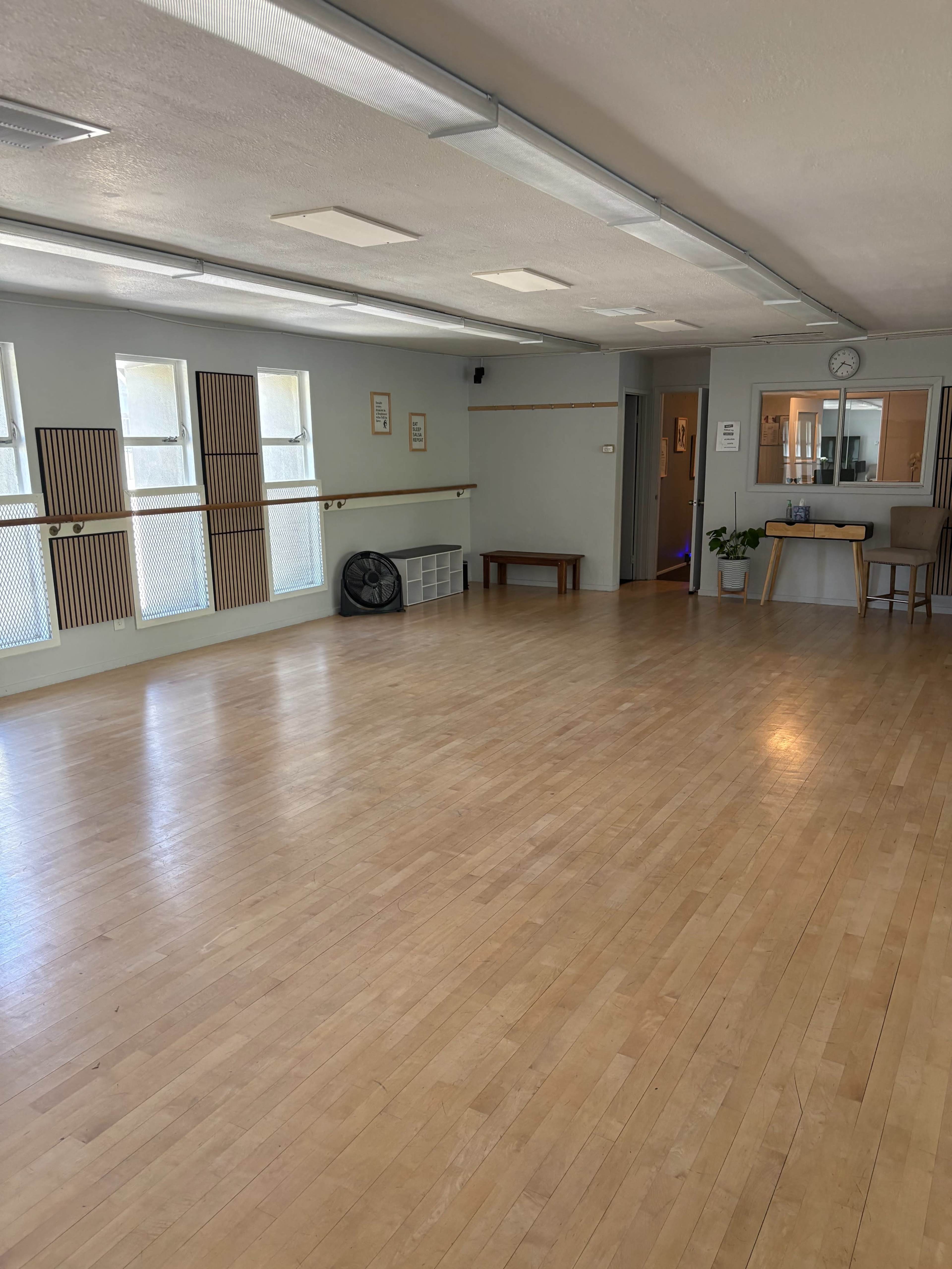 The image shows a spacious, empty room with wooden flooring, large windows, and a small kitchenette area at one end.