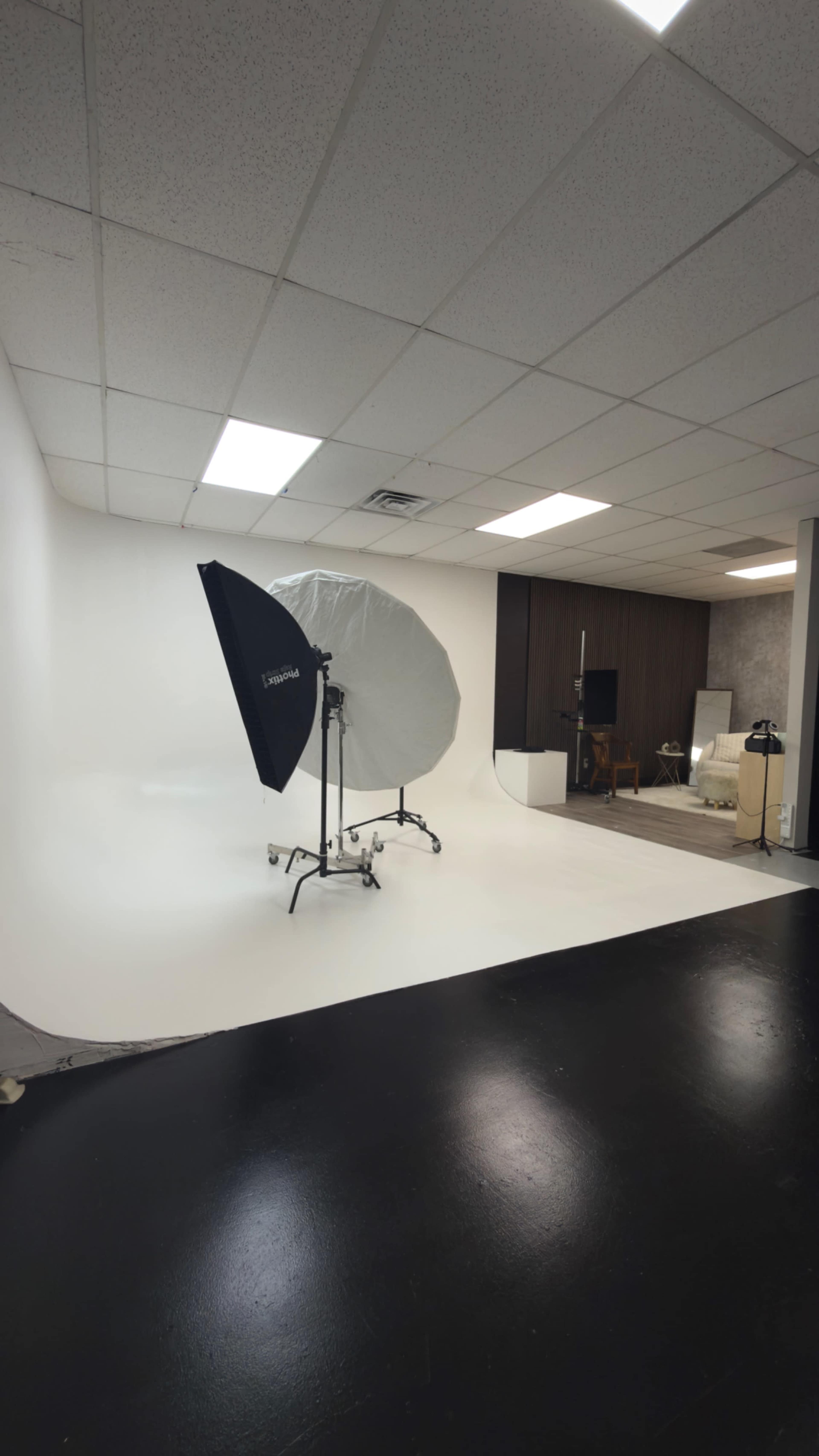The image shows a photography studio featuring a large softbox umbrella on a stand, white backdrop, and a section of dark flooring.