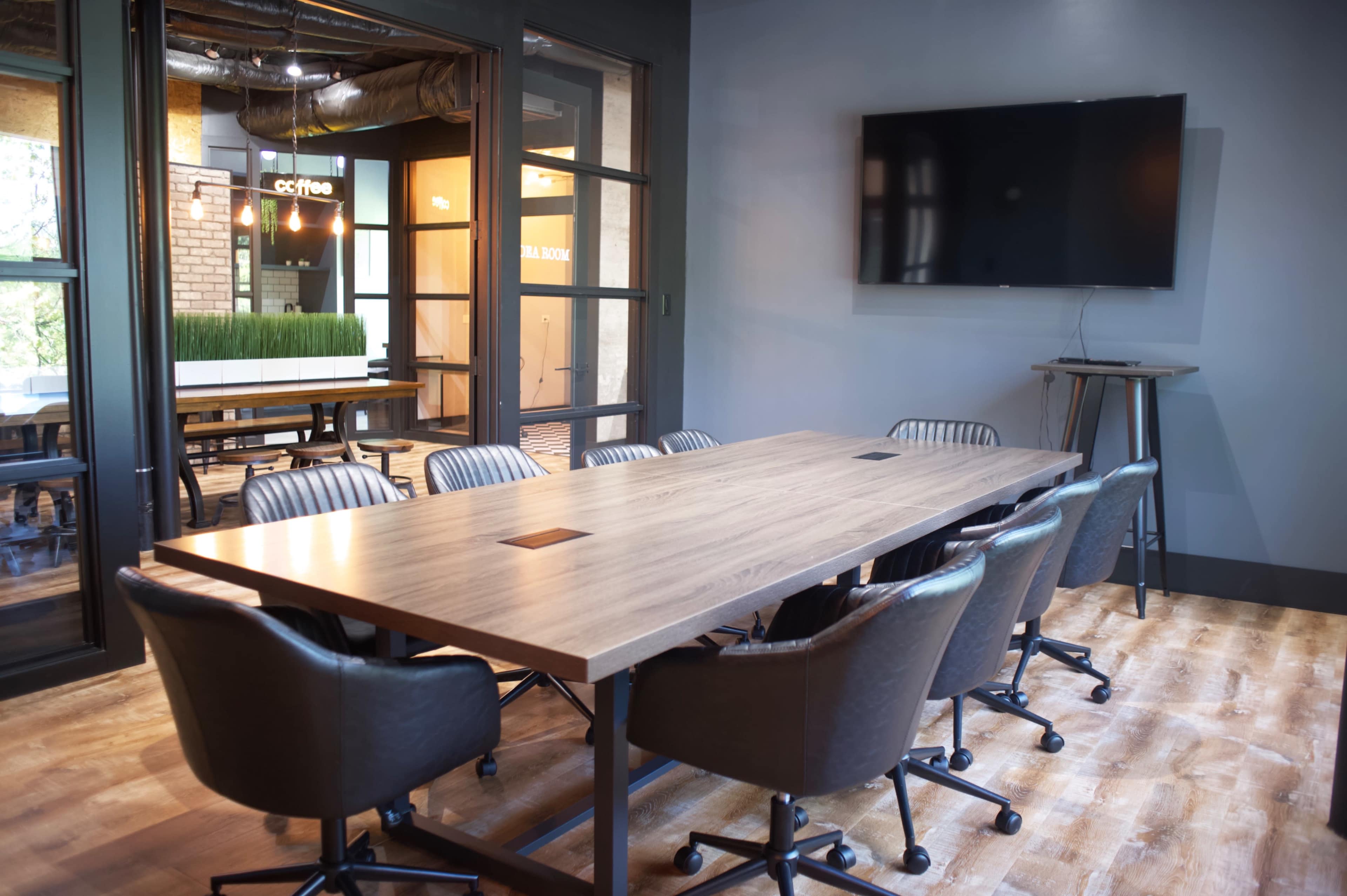 A modern conference room features a large wooden table surrounded by ergonomic chairs and a wall-mounted television.