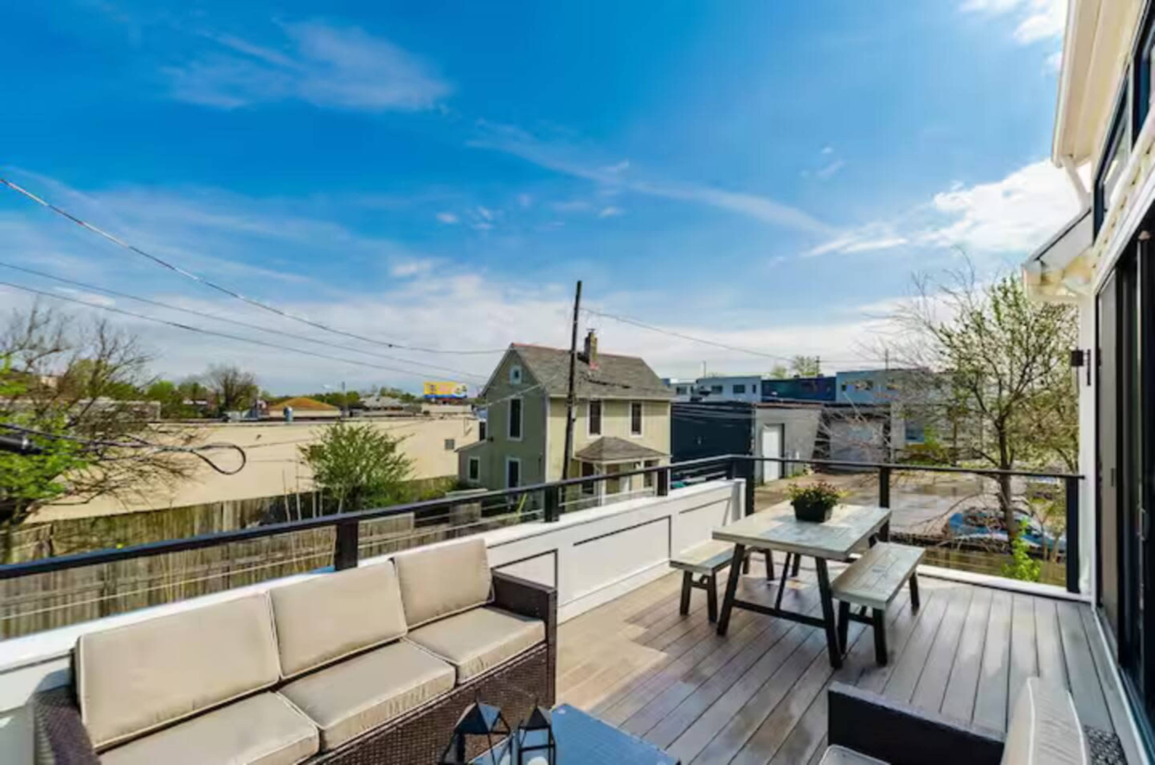 A modern rooftop terrace features seating, a table, and a view of nearby buildings and trees against a clear blue sky.