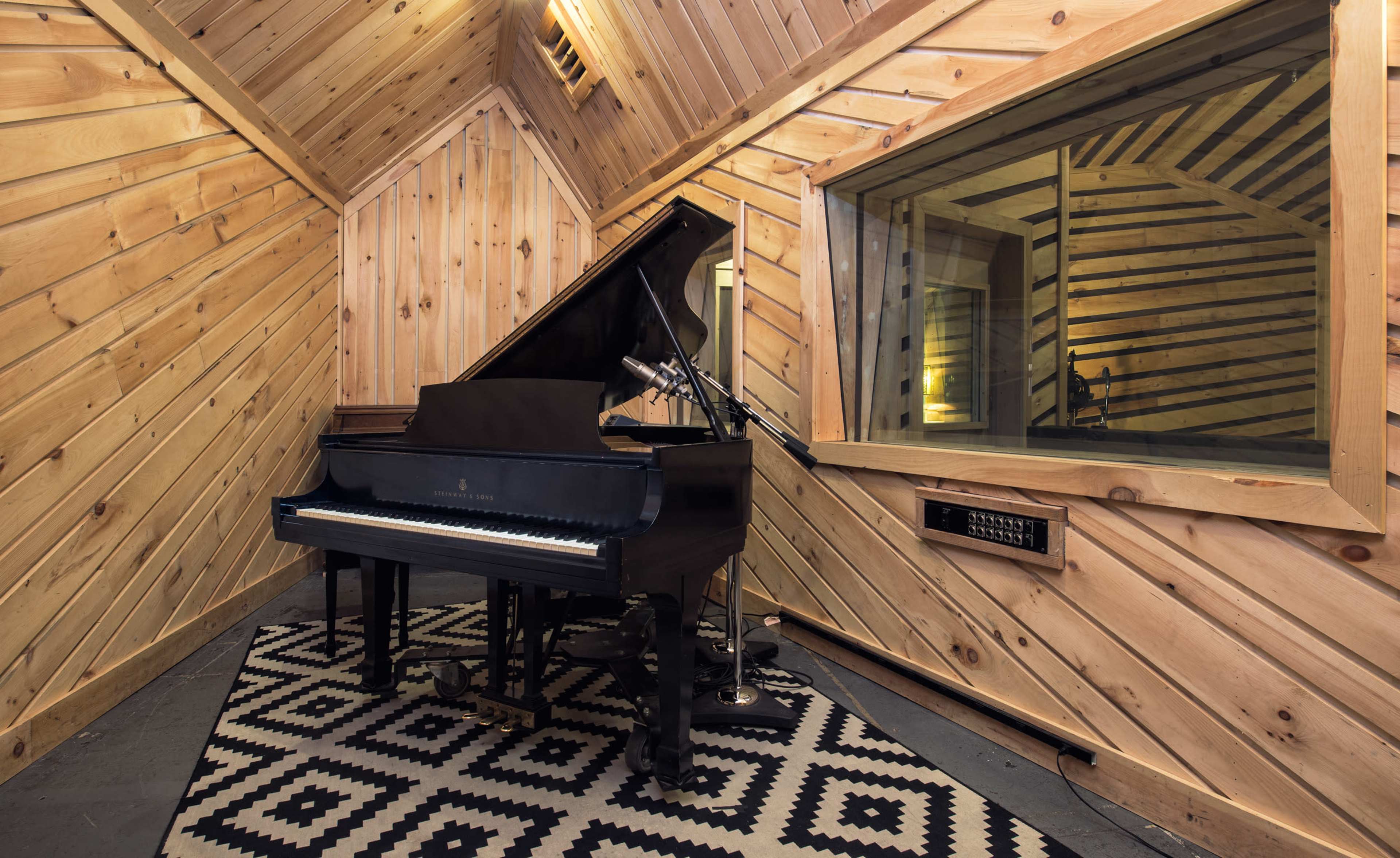 A black grand piano is positioned in a wooden room with diagonal paneling and a large window overlooking another space.