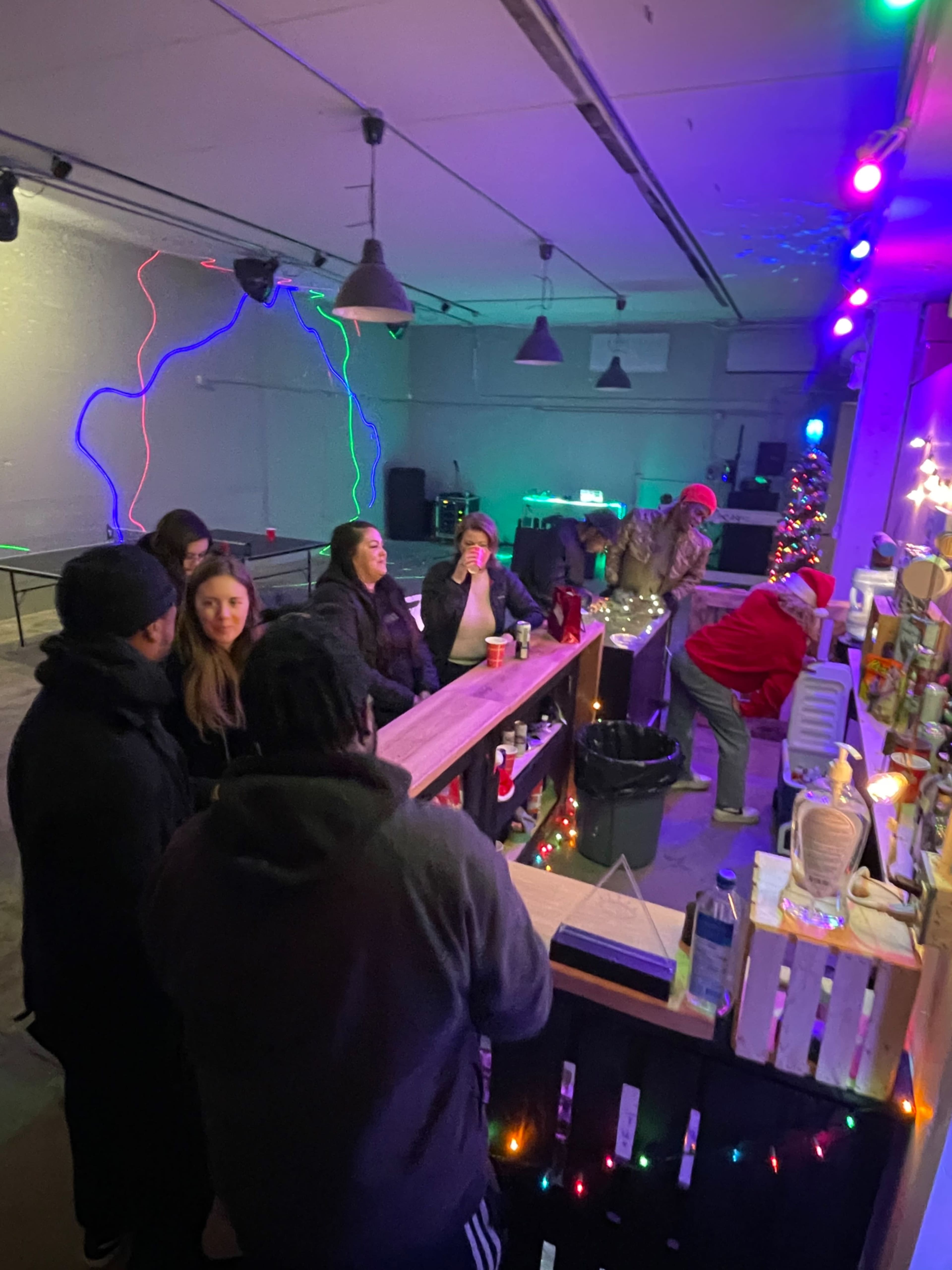 A group of people gather at a bar area decorated with colorful lighting and festive decorations.