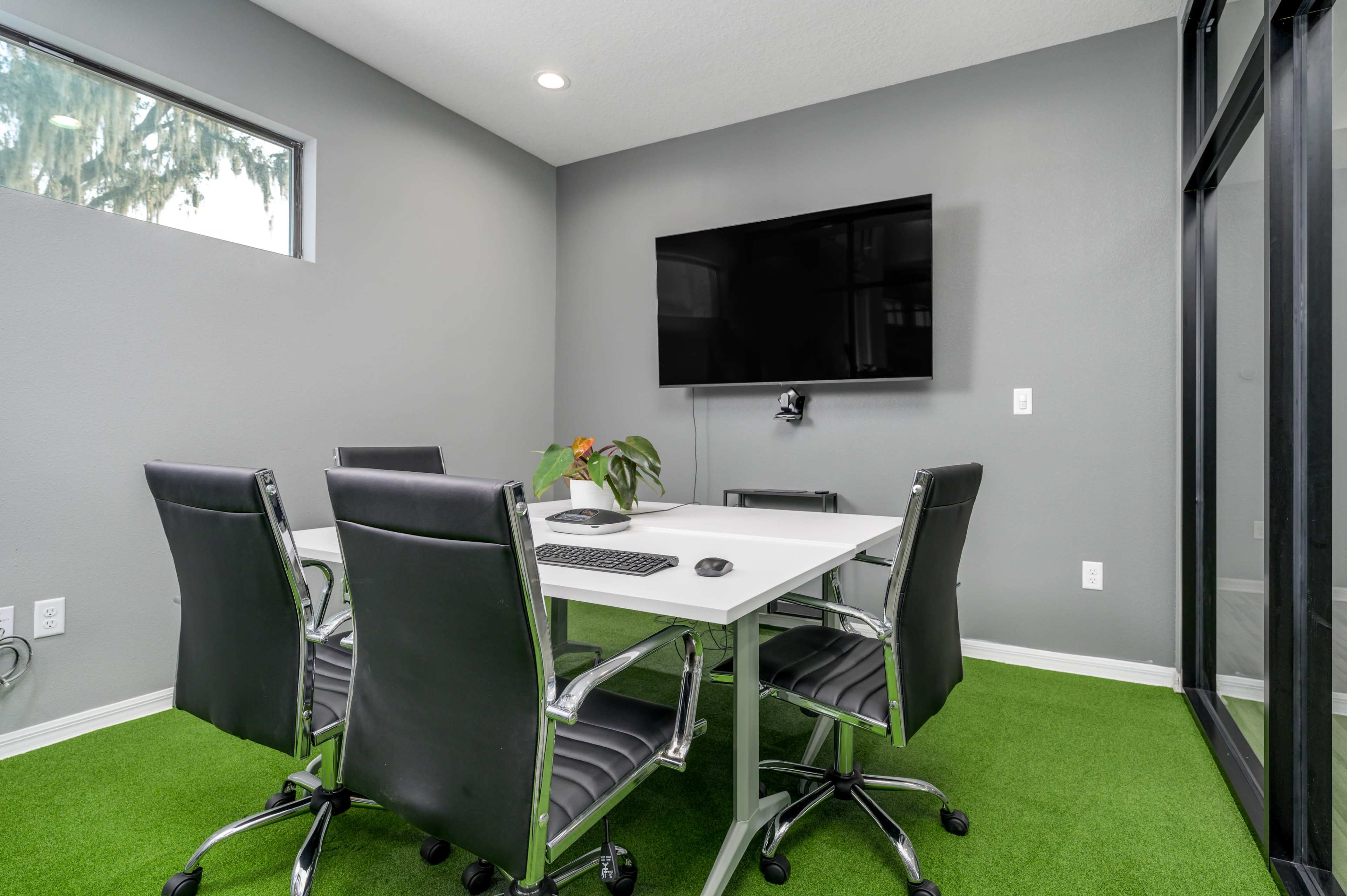 A conference room with a white table, black chairs, a green carpet, and a mounted television on a gray wall.