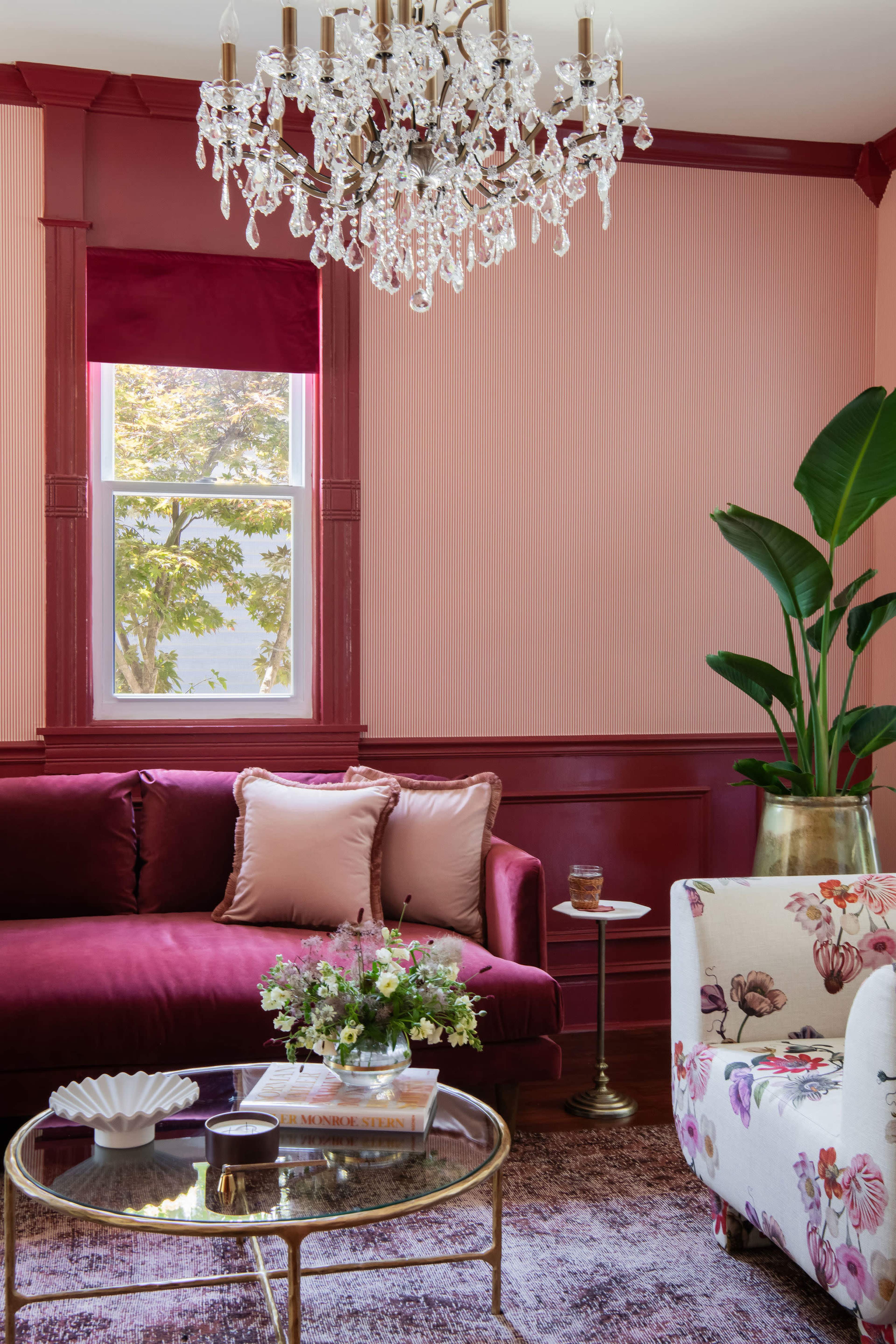 The image shows a stylish living room featuring a burgundy sofa, a floral accent chair, a glass coffee table, and a crystal chandelier above.