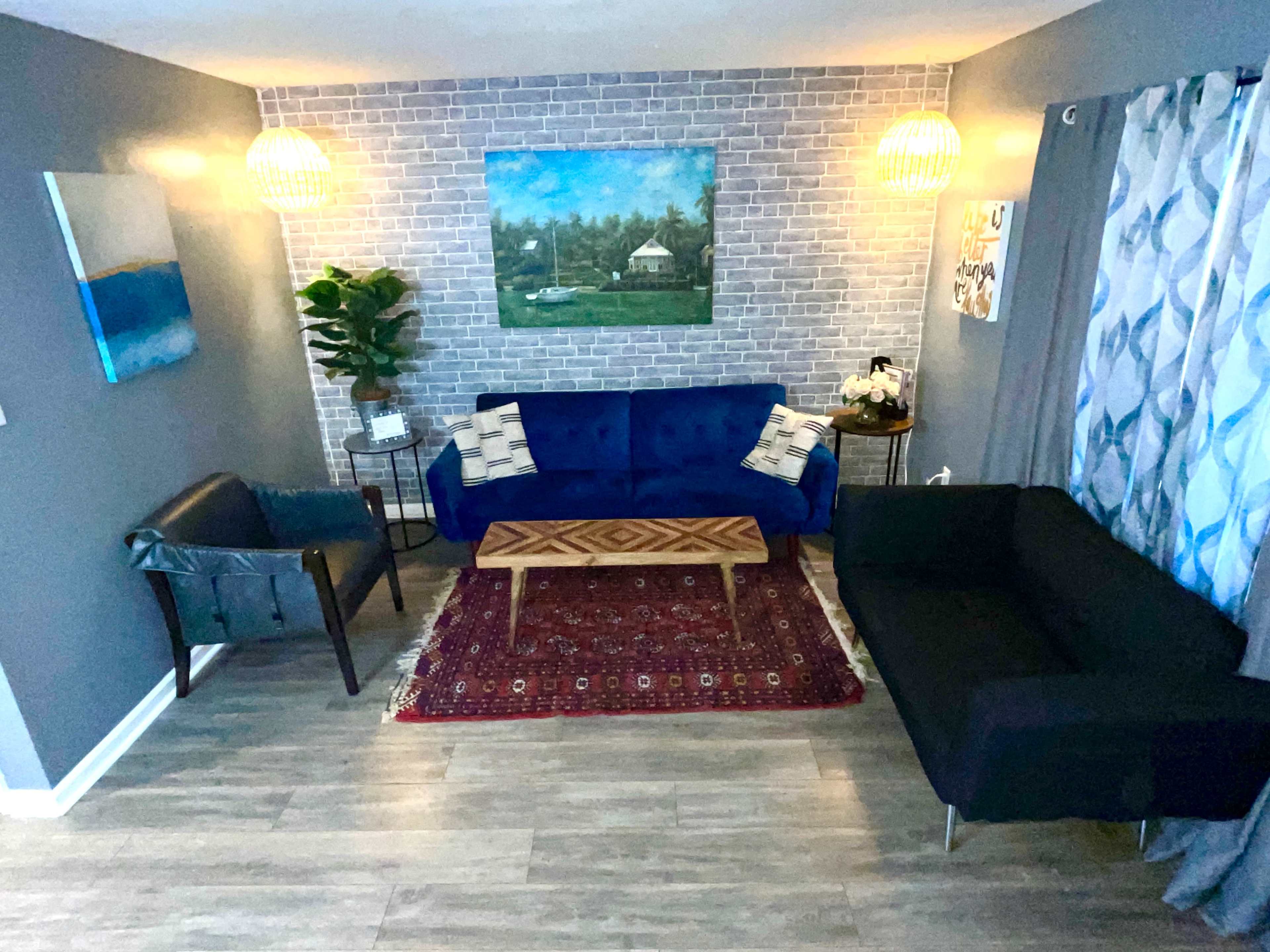 The image shows a cozy living room with a brick accent wall, two sofas facing each other, a wooden coffee table in the center, and decorative elements like plants and artwork.