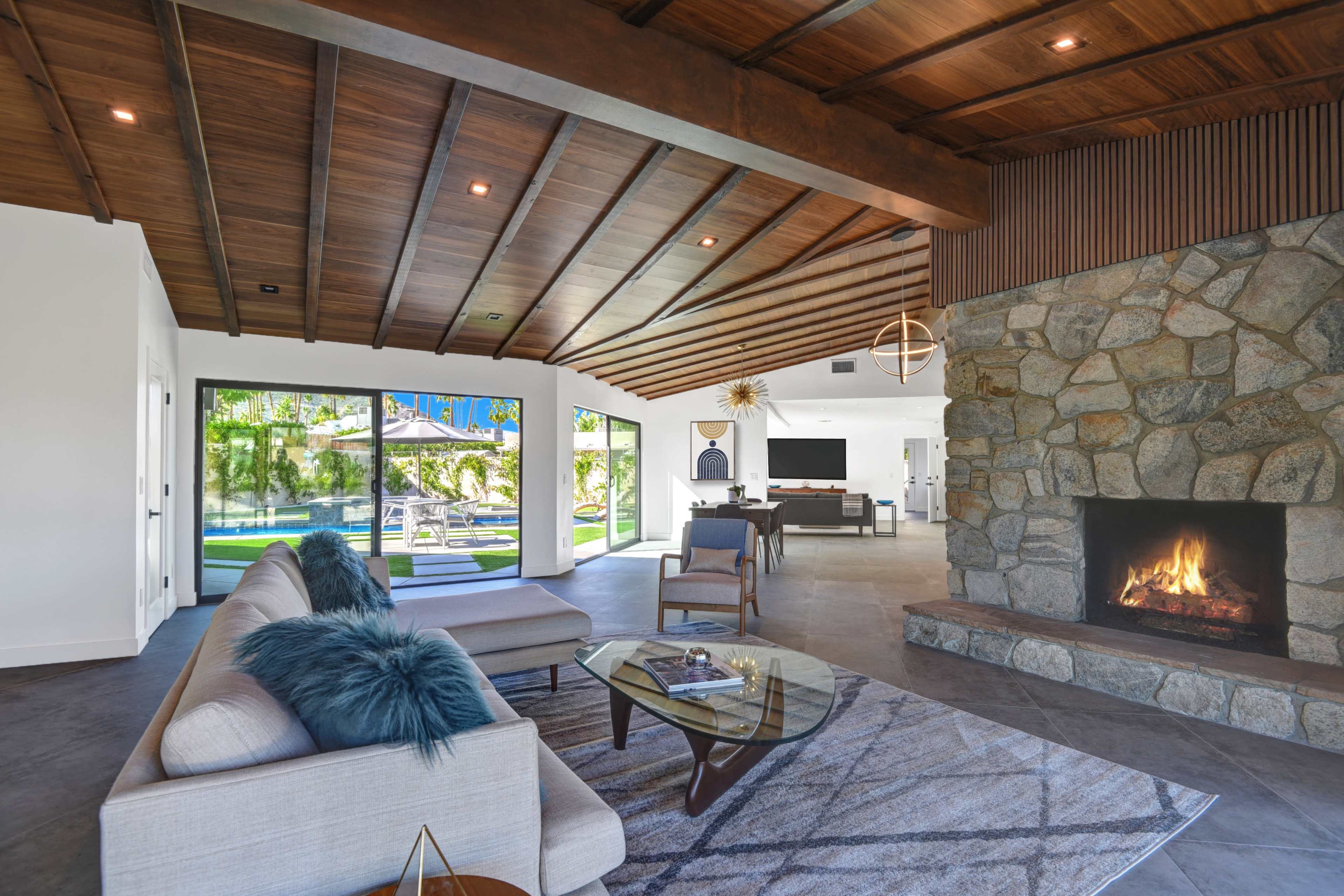 A modern living room features a stone fireplace, wooden ceiling beams, and large glass doors opening to an outdoor space.