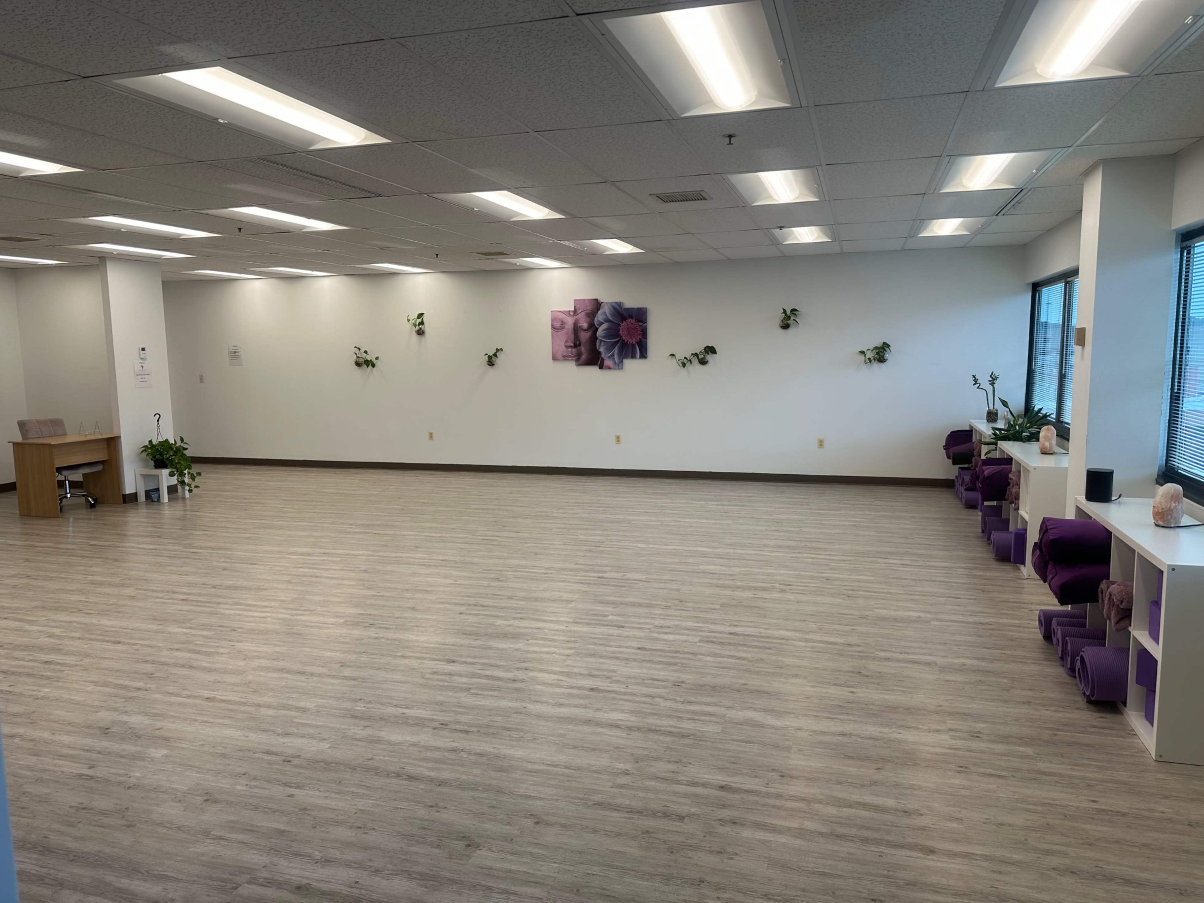 A spacious, empty room with light wood flooring, purple seating against the wall, and plants along the edges.