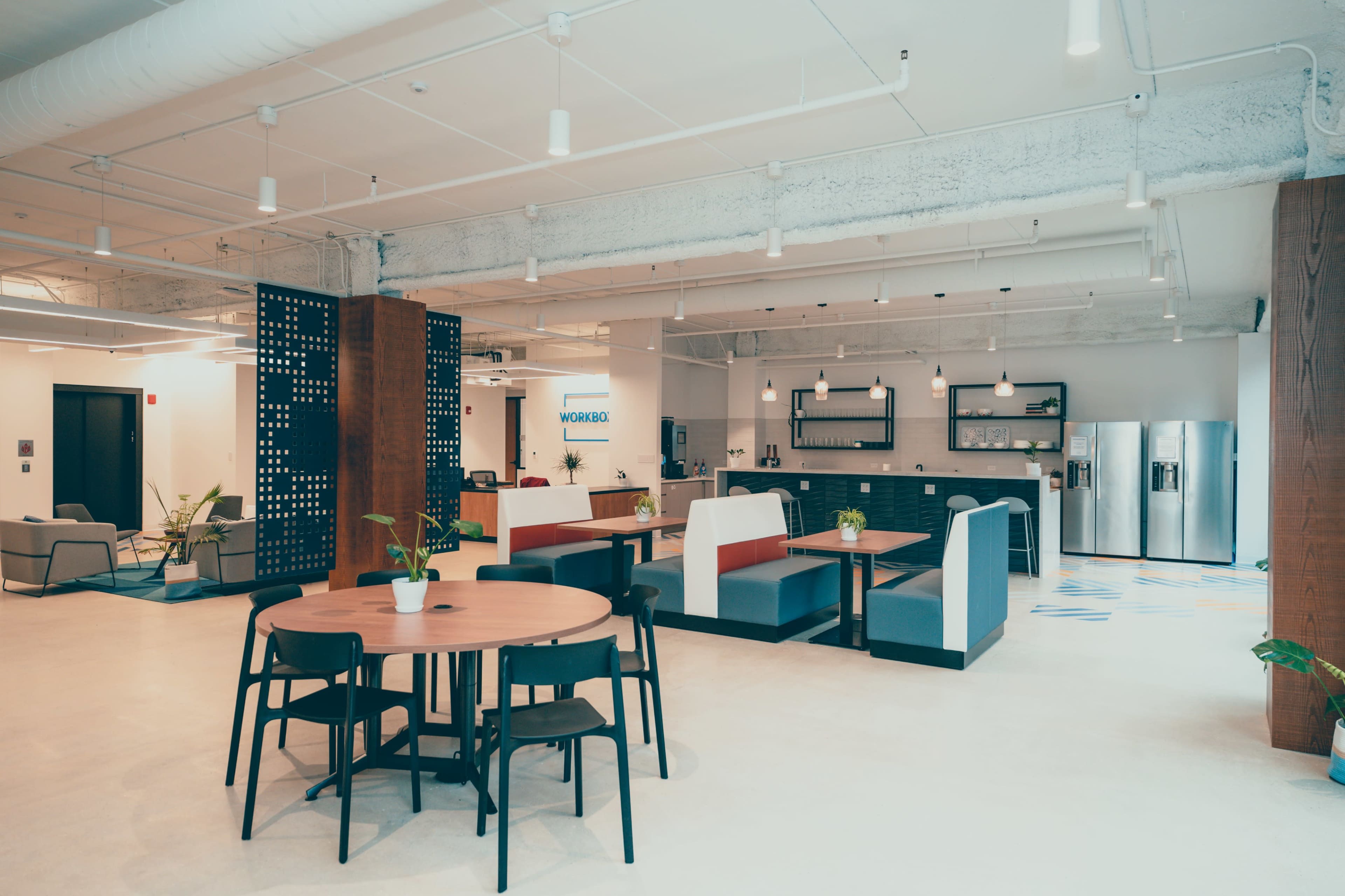 The image shows a modern office space featuring a kitchen area, seating booths, and a dining table surrounded by black chairs.