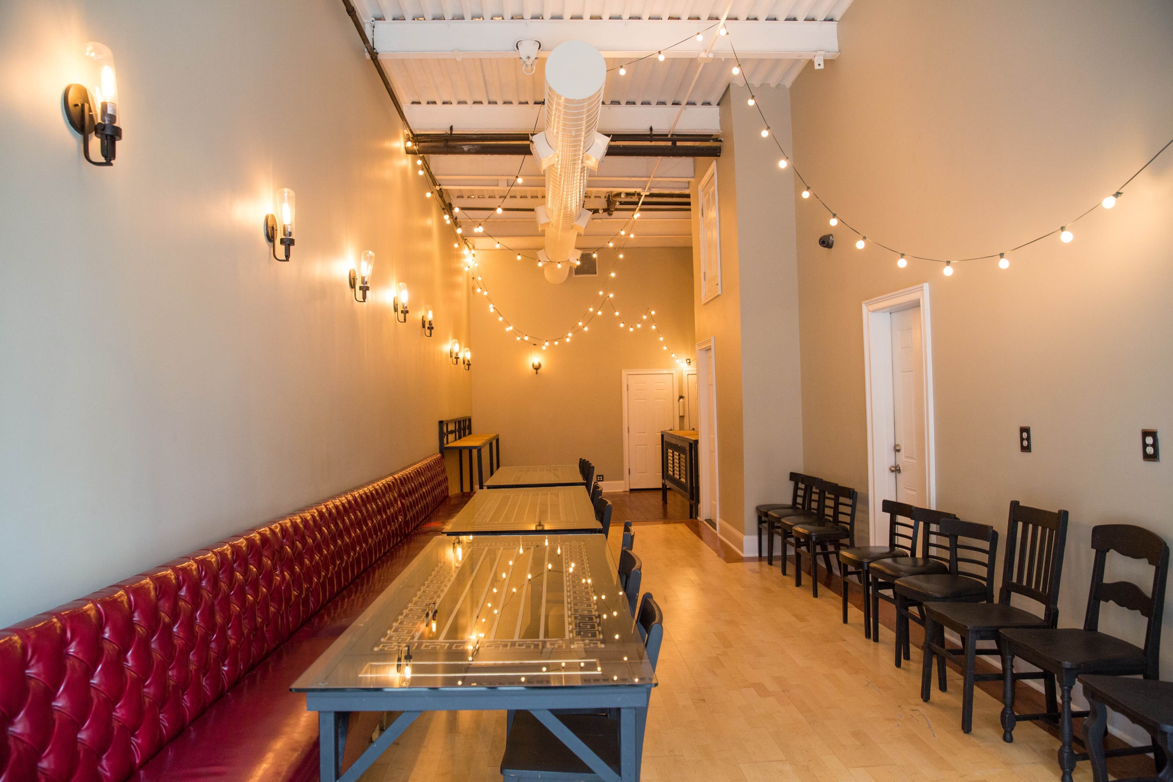 The image shows a long, narrow room with red tufted seating on one side, wooden tables in the center, and string lights hanging from the ceiling.