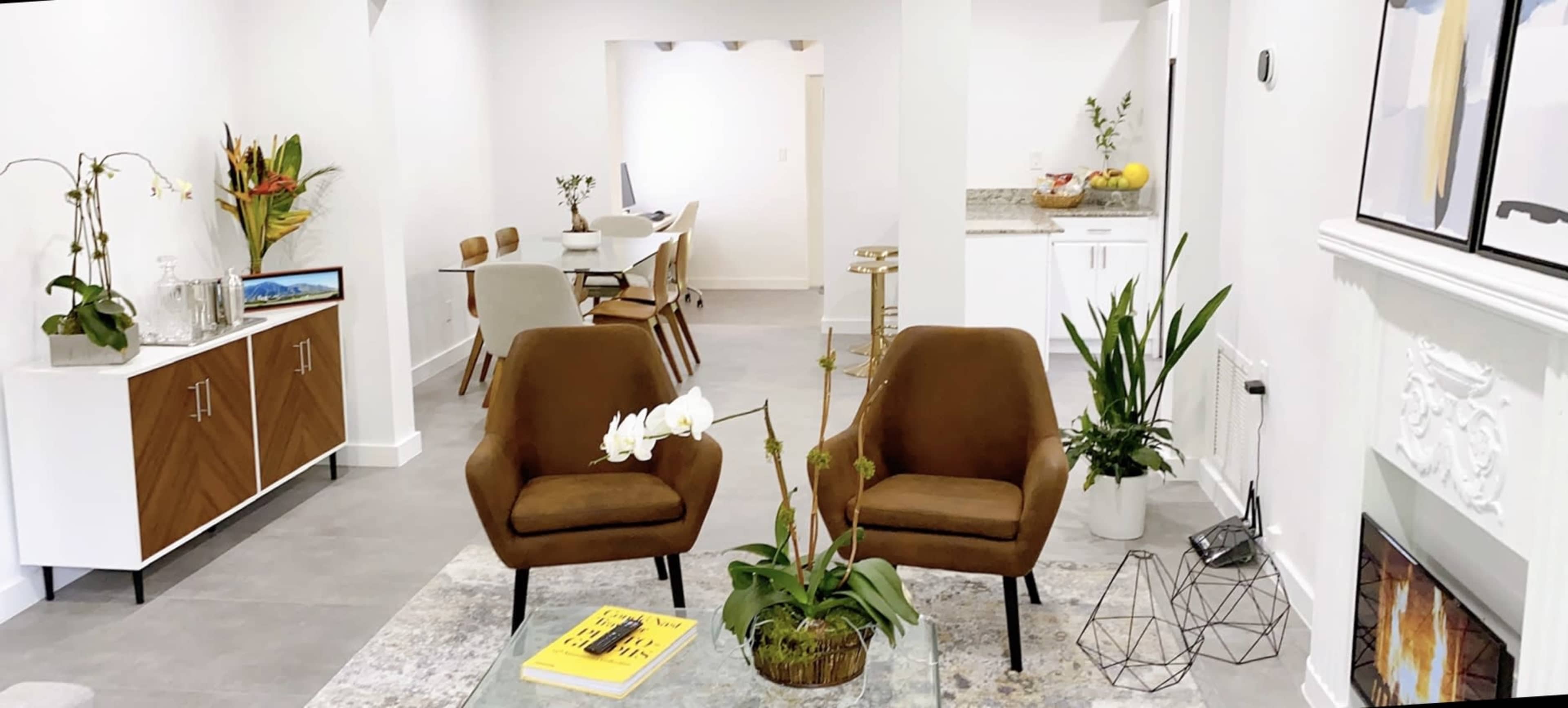 The image shows a modern interior space featuring two brown armchairs on a rug, with a dining area and kitchen visible in the background.