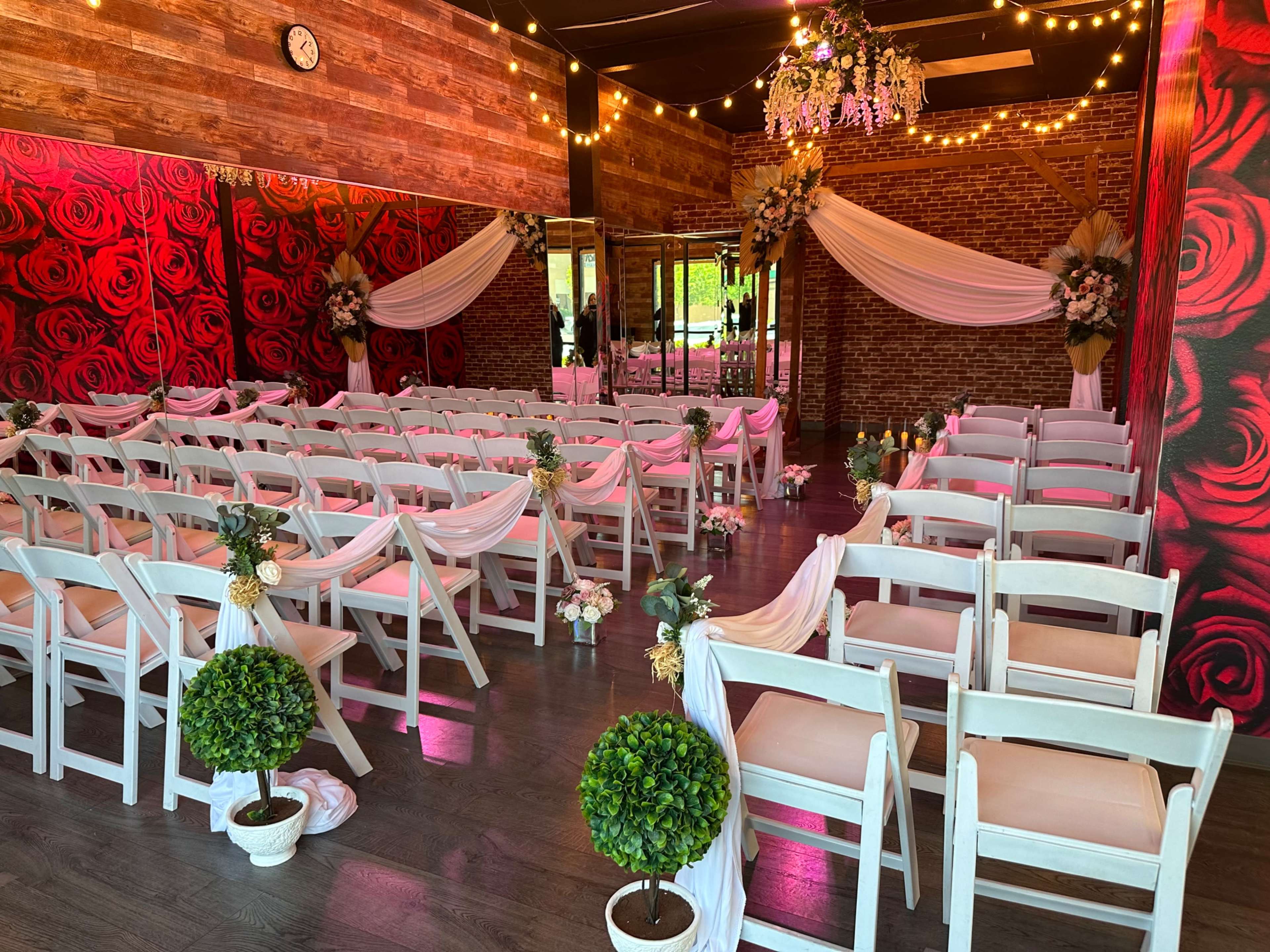 A decorated event space features rows of white chairs with floral accents, arranged facing a small altar, surrounded by rose-themed decor and soft lighting.