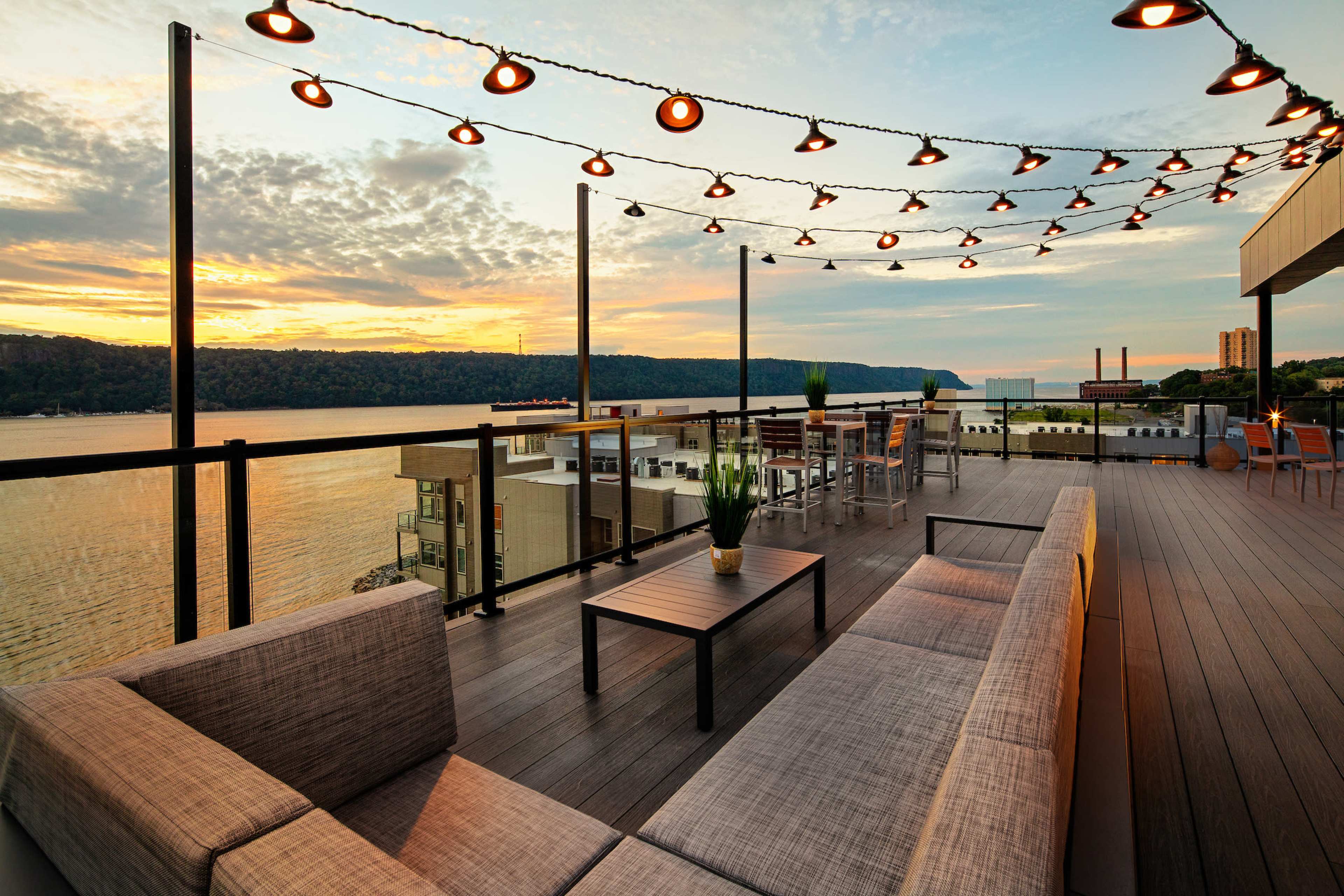 The image shows a modern outdoor terrace with a sectional sofa, a wooden table, and string lights, overlooking a river at sunset.