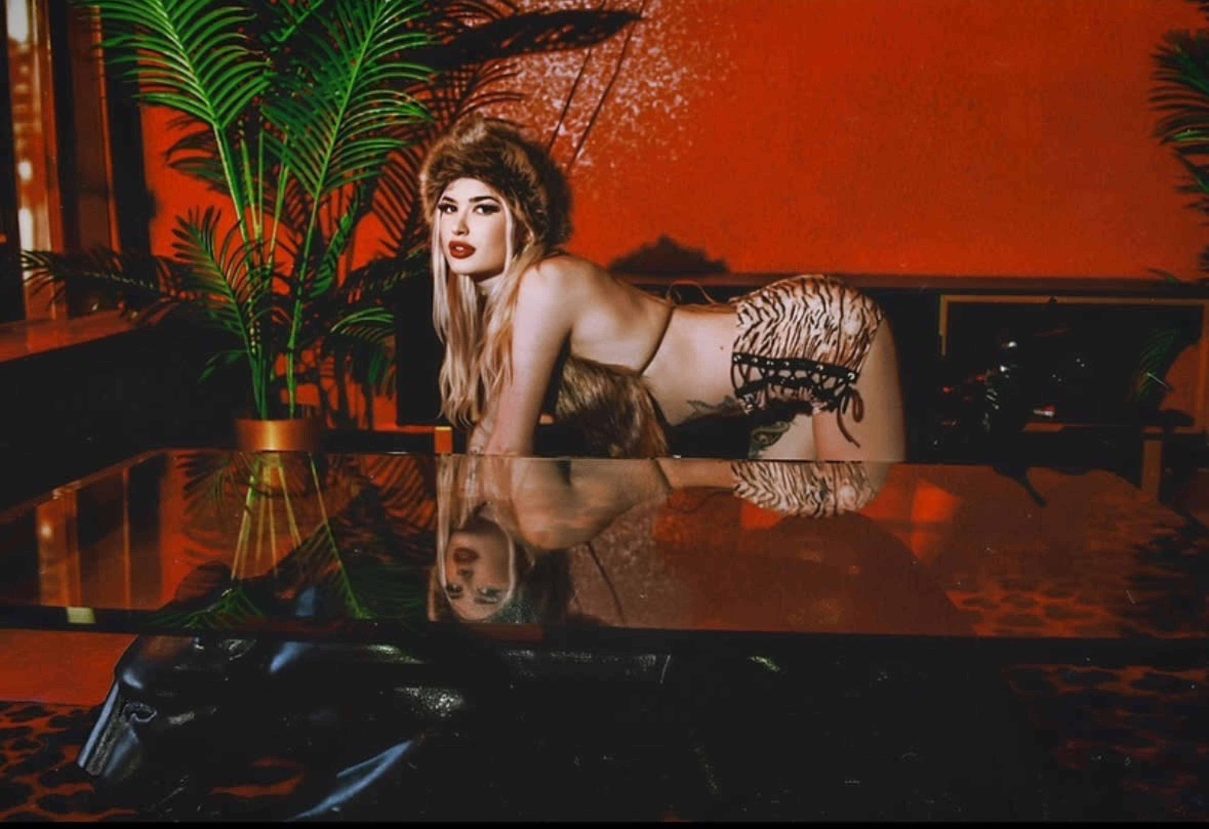 A person in a fur hat poses on all fours on a table in a room with red walls and decorative plants.