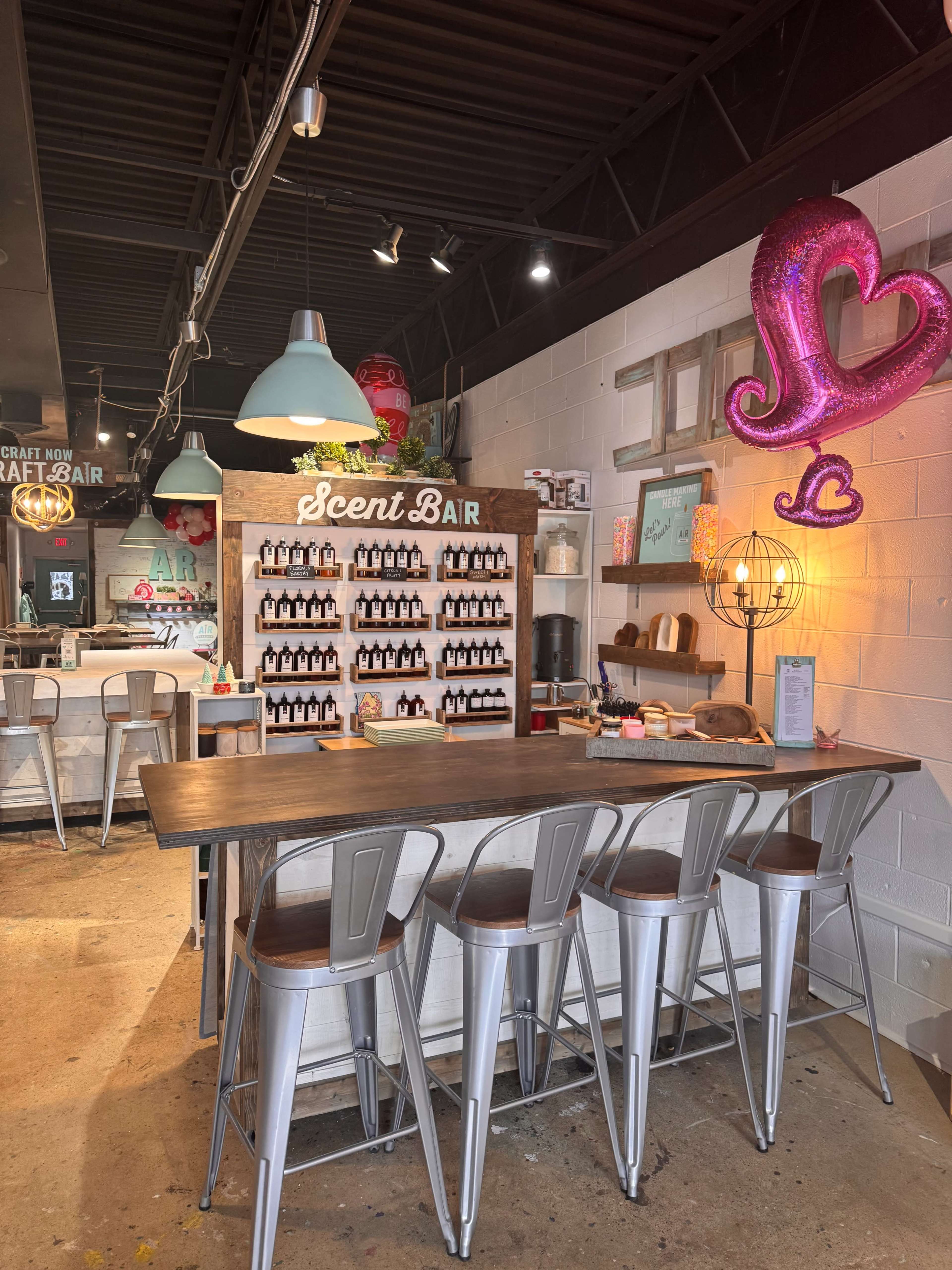 The image shows a well-arranged scent bar featuring various bottles on a wooden shelf, accompanied by metal stools and decorative elements in a modern interior space.