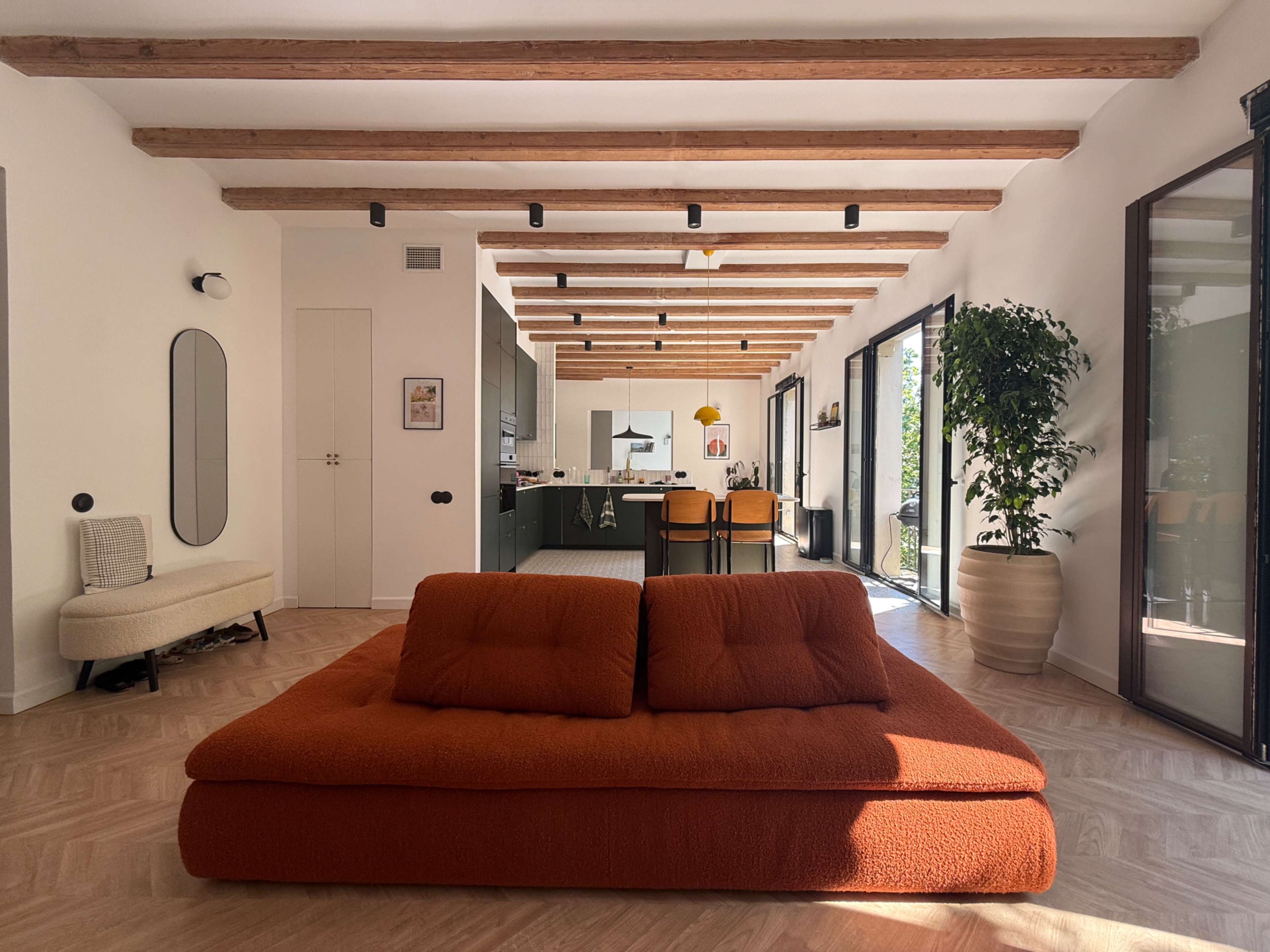 The image shows a modern living space with a large orange sofa in the center, wooden beams on the ceiling, and an open kitchen area in the background.
