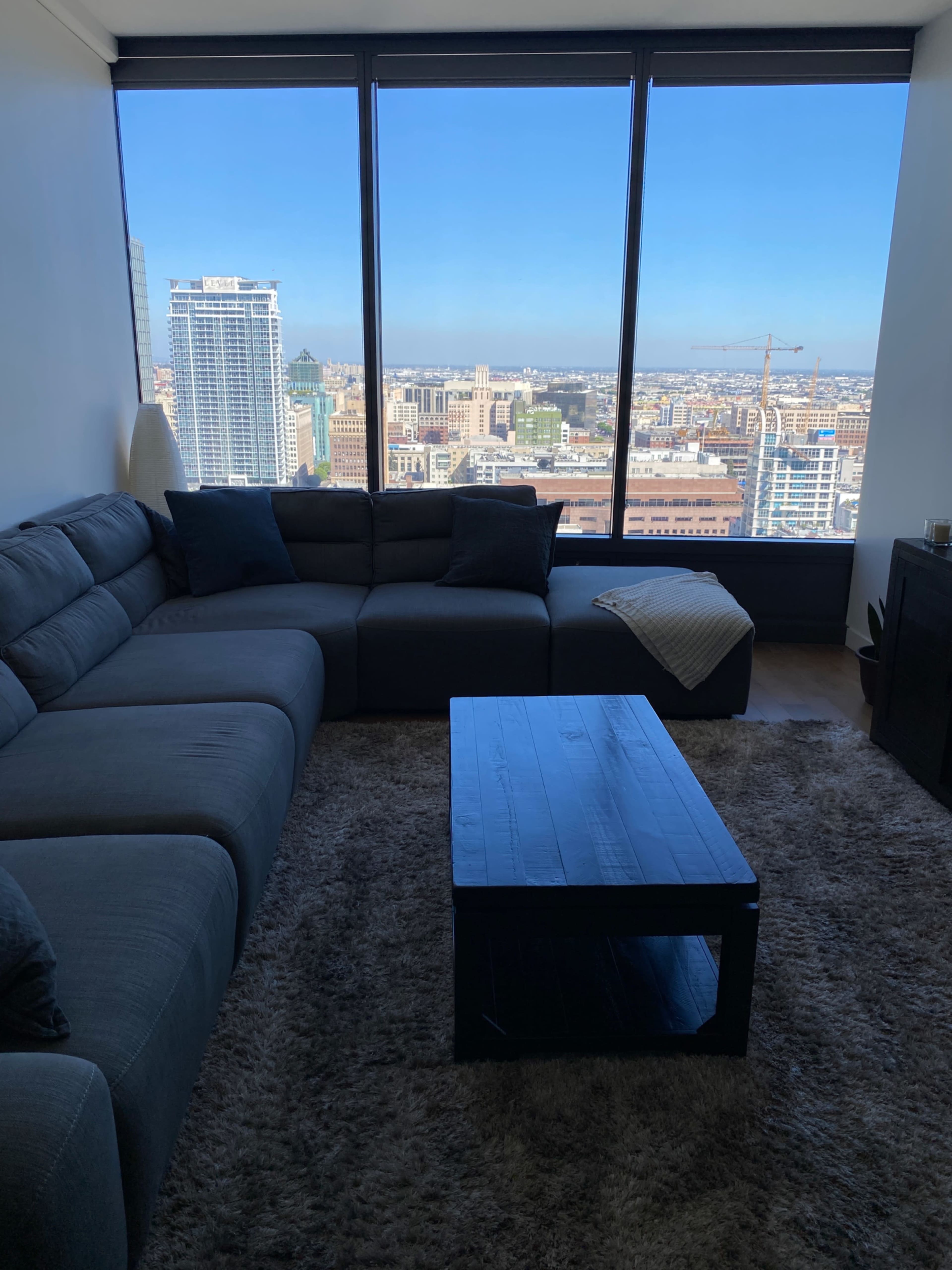 A modern living room features a gray sectional sofa and a black coffee table with a city skyline visible through large floor-to-ceiling windows.