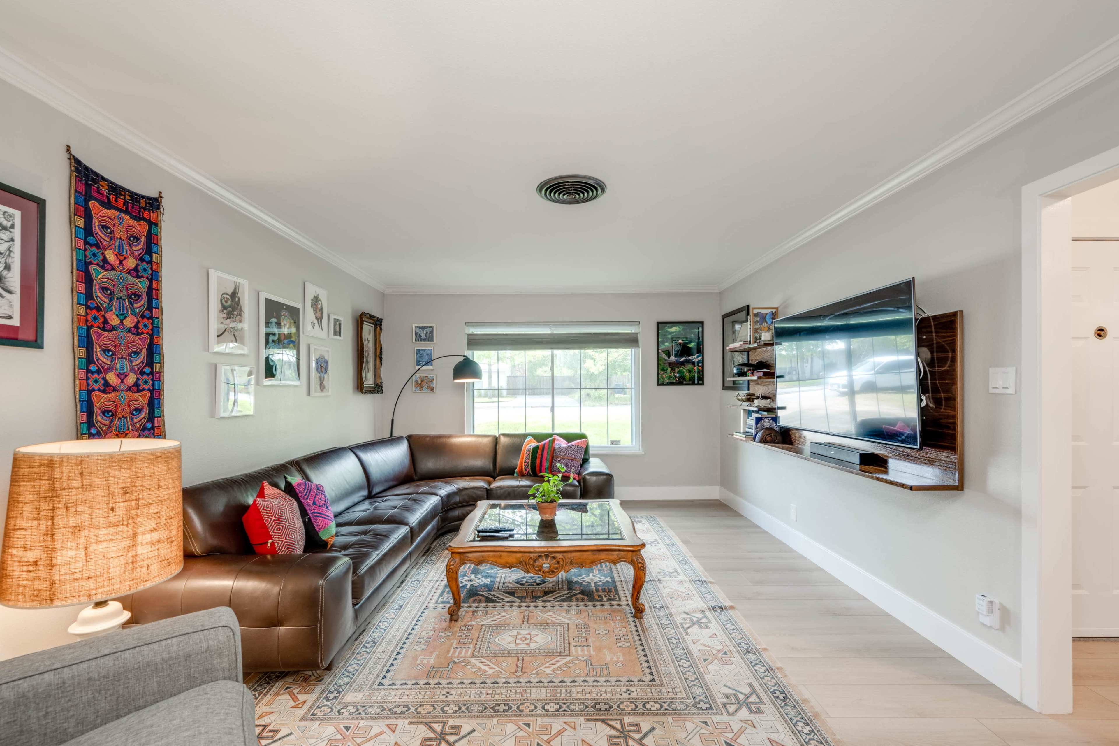 A spacious living room features a leather sectional sofa, a wooden coffee table, a flat-screen TV mounted on the wall, and large windows allowing natural light to enter.