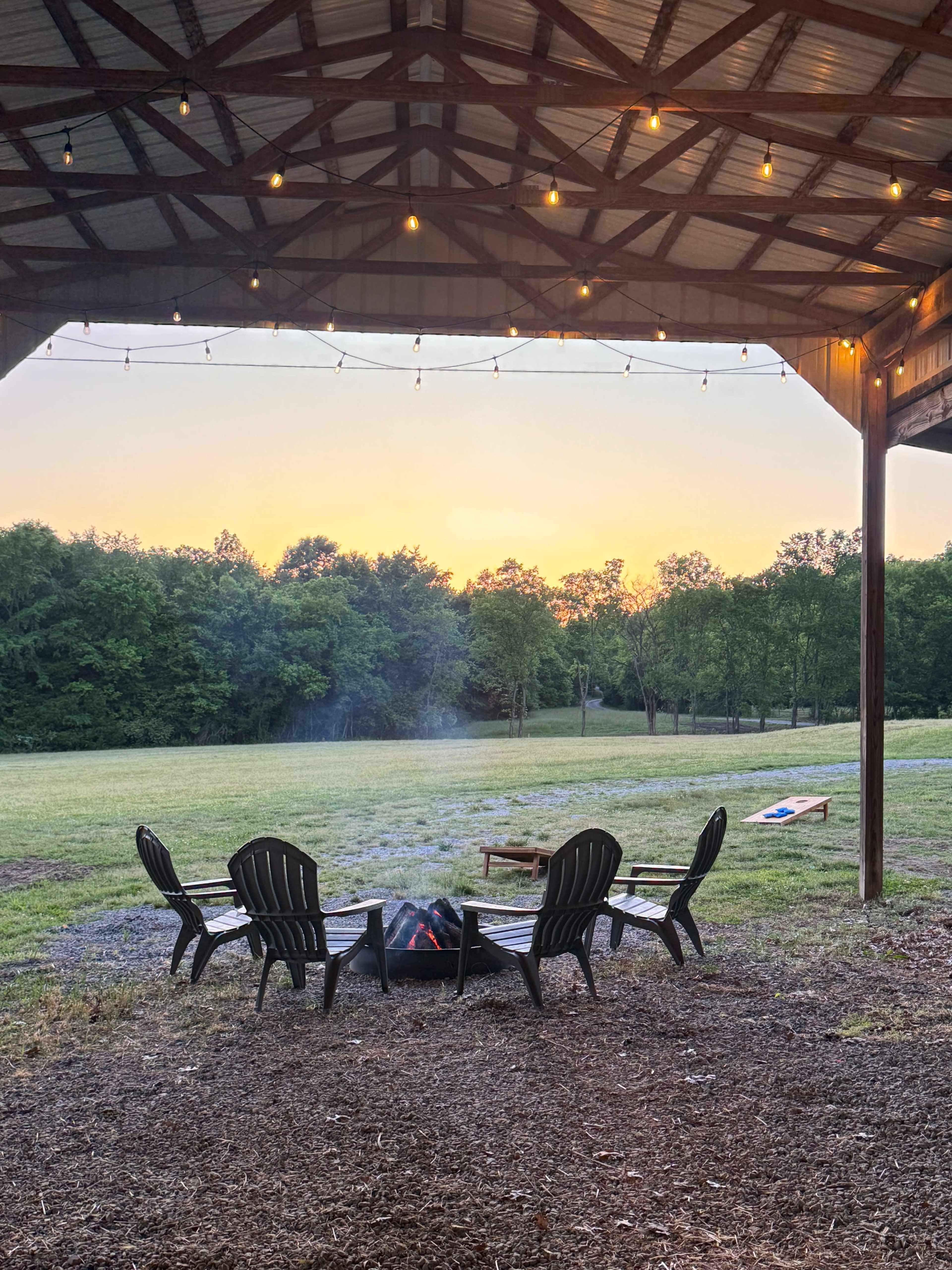A circle of black Adirondack chairs surrounds a fire pit under a covered wooden structure, with a sunset lighting up the sky in the background.