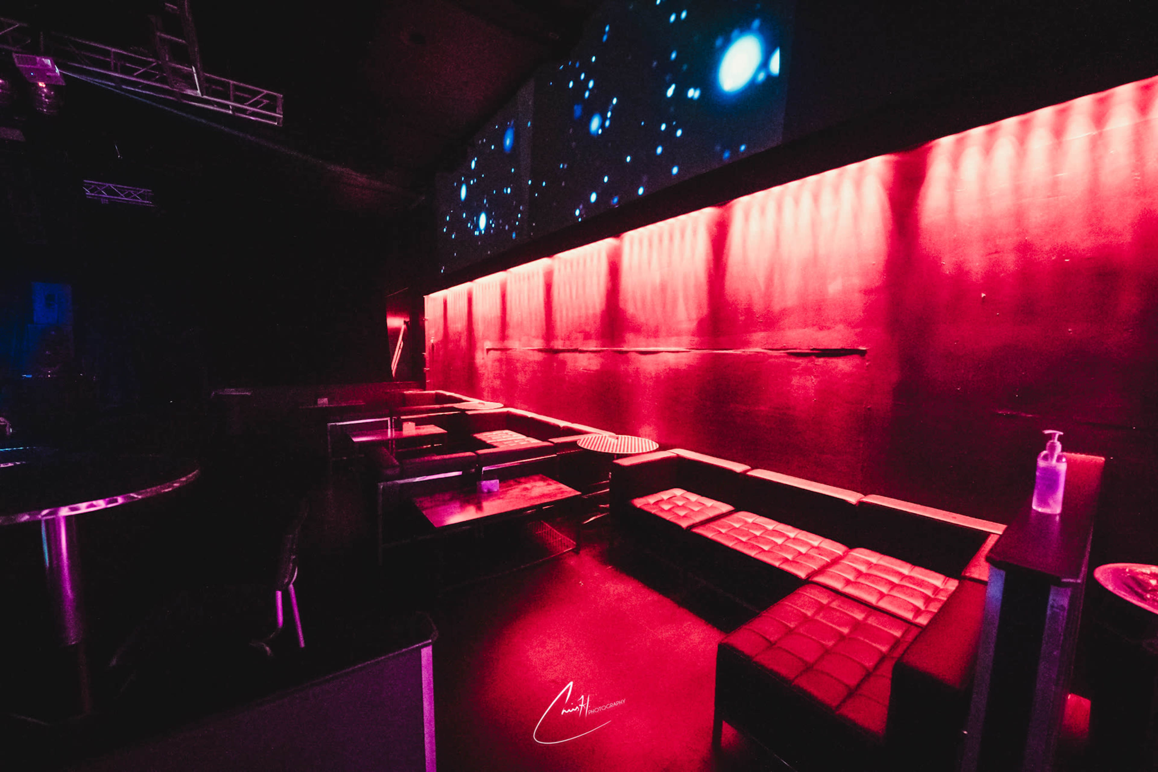 The image shows a dimly lit bar or lounge area with red lighting, featuring modern seating and a projector screen displaying abstract visuals.
