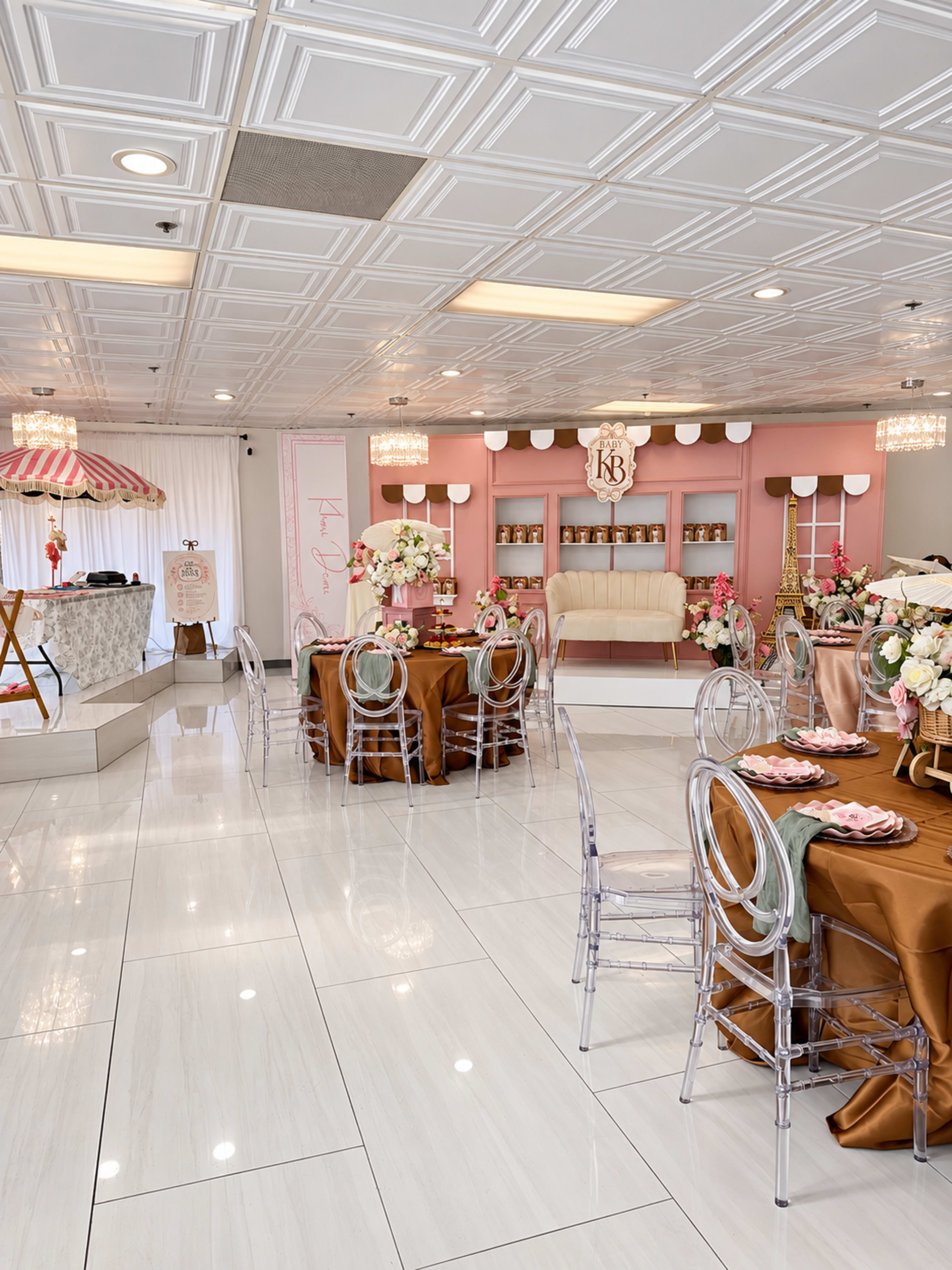 The image shows a decorated event space with tables set for a celebration, featuring pink and gold accents, floral arrangements, and a dessert display.