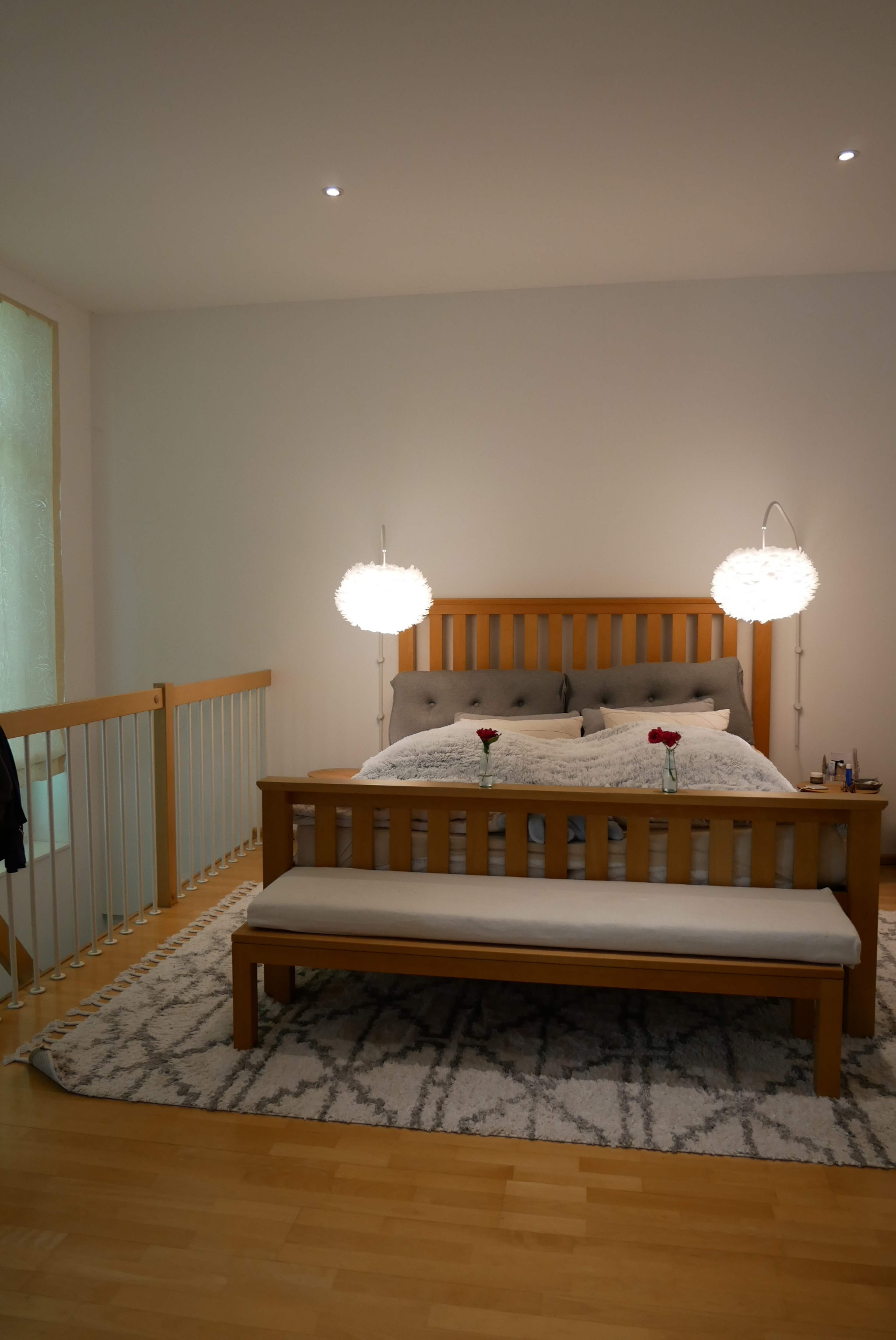 A wooden bed with decorative pillows and roses sits in a softly lit bedroom, accompanied by a bench and two hanging lights.