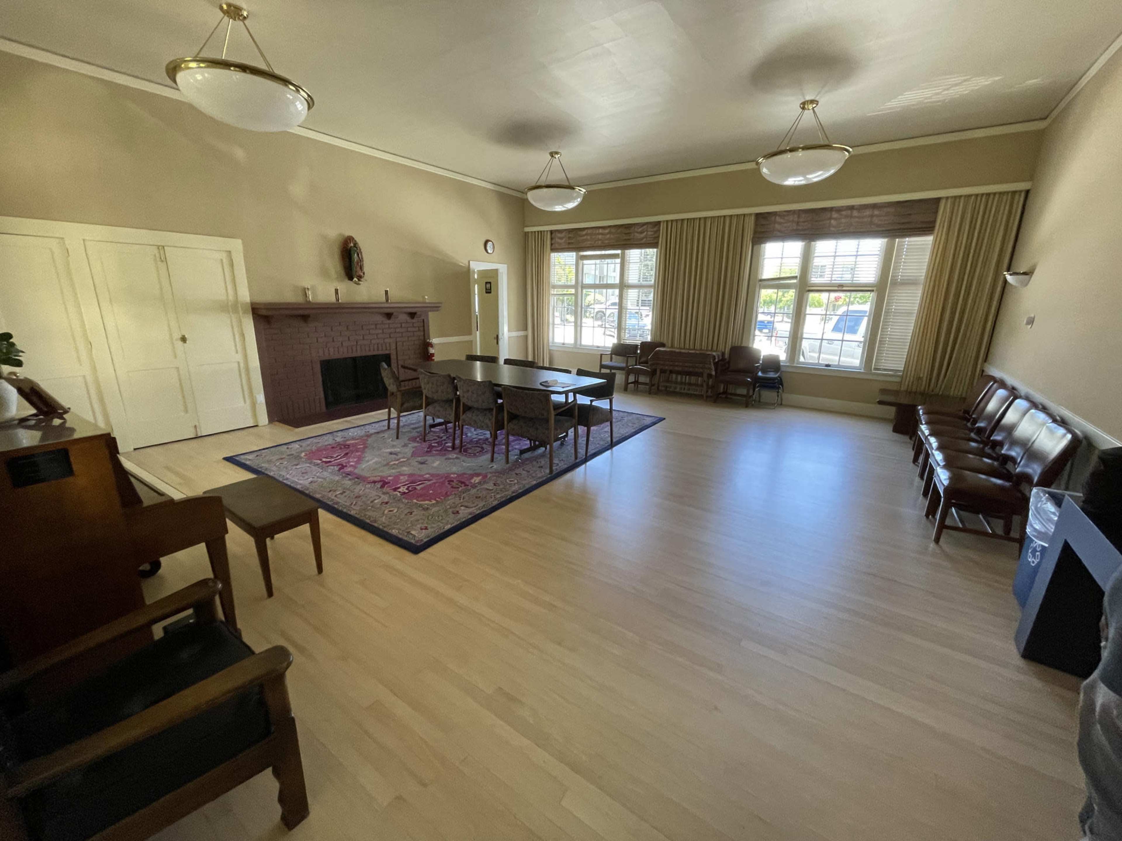 The room features wooden flooring, a large rug in the center, a long table surrounded by chairs, and a fireplace against the wall.