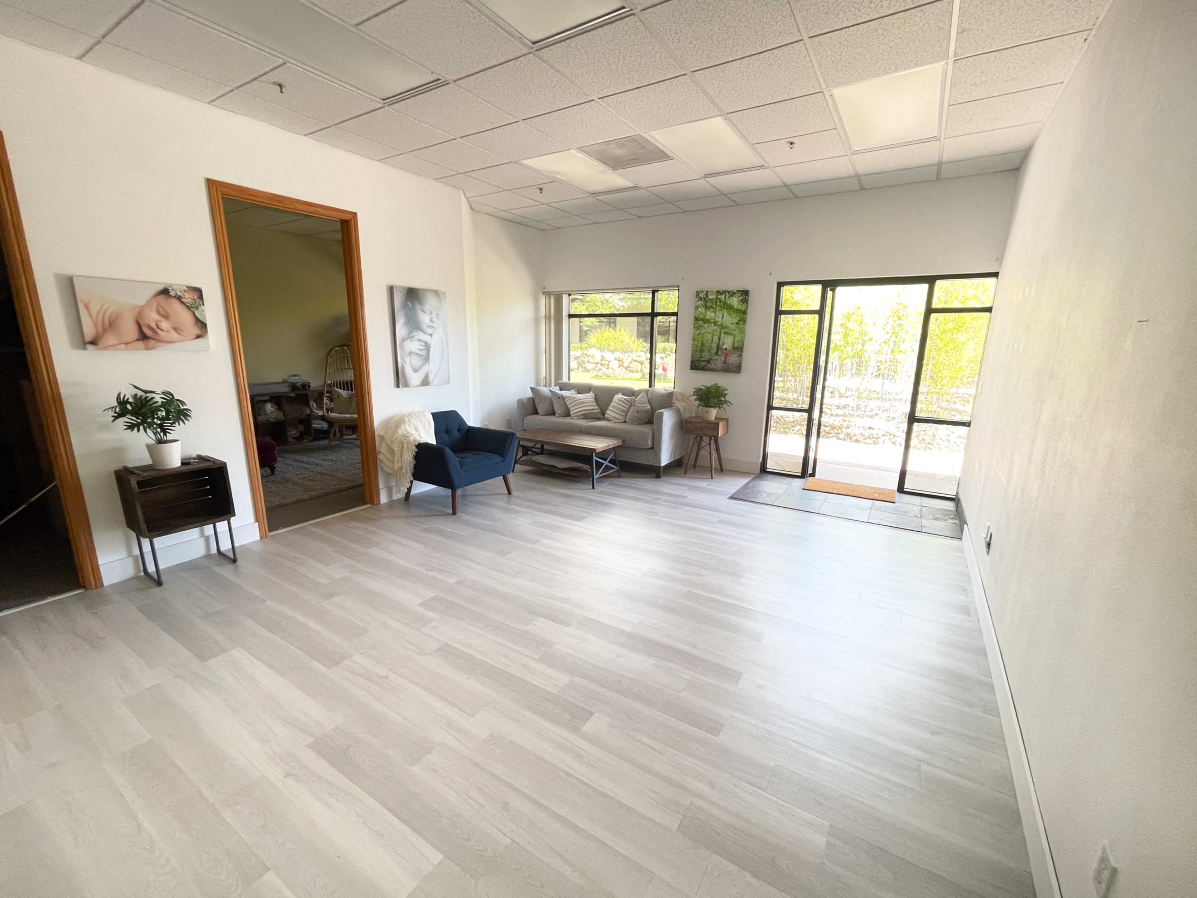 The image shows a spacious, bright room with light-colored wooden flooring, featuring seating arrangements, a plant, and large windows that provide natural light.