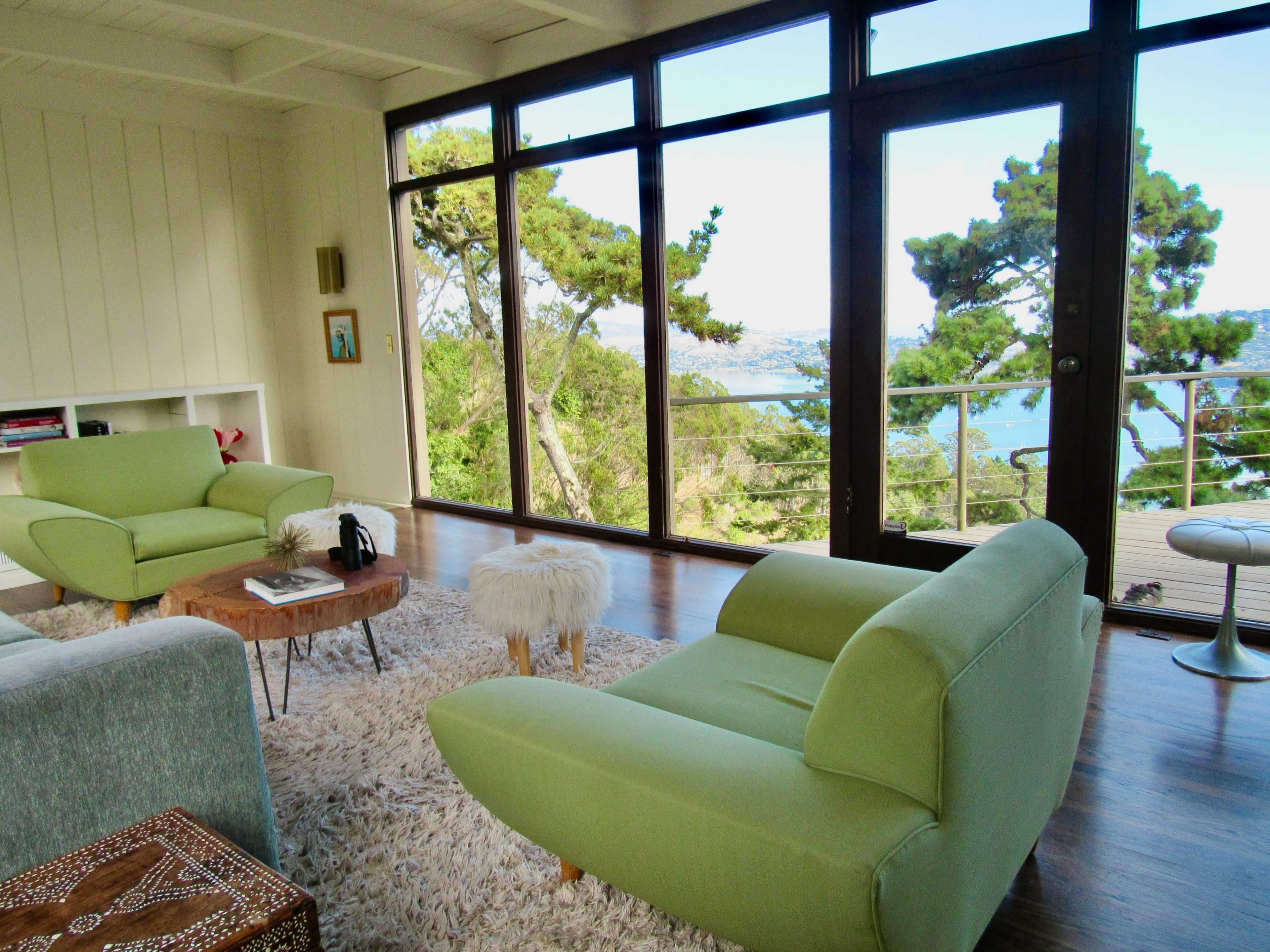 A brightly lit room with two green chairs, a coffee table, and large windows offering a view of trees and water.